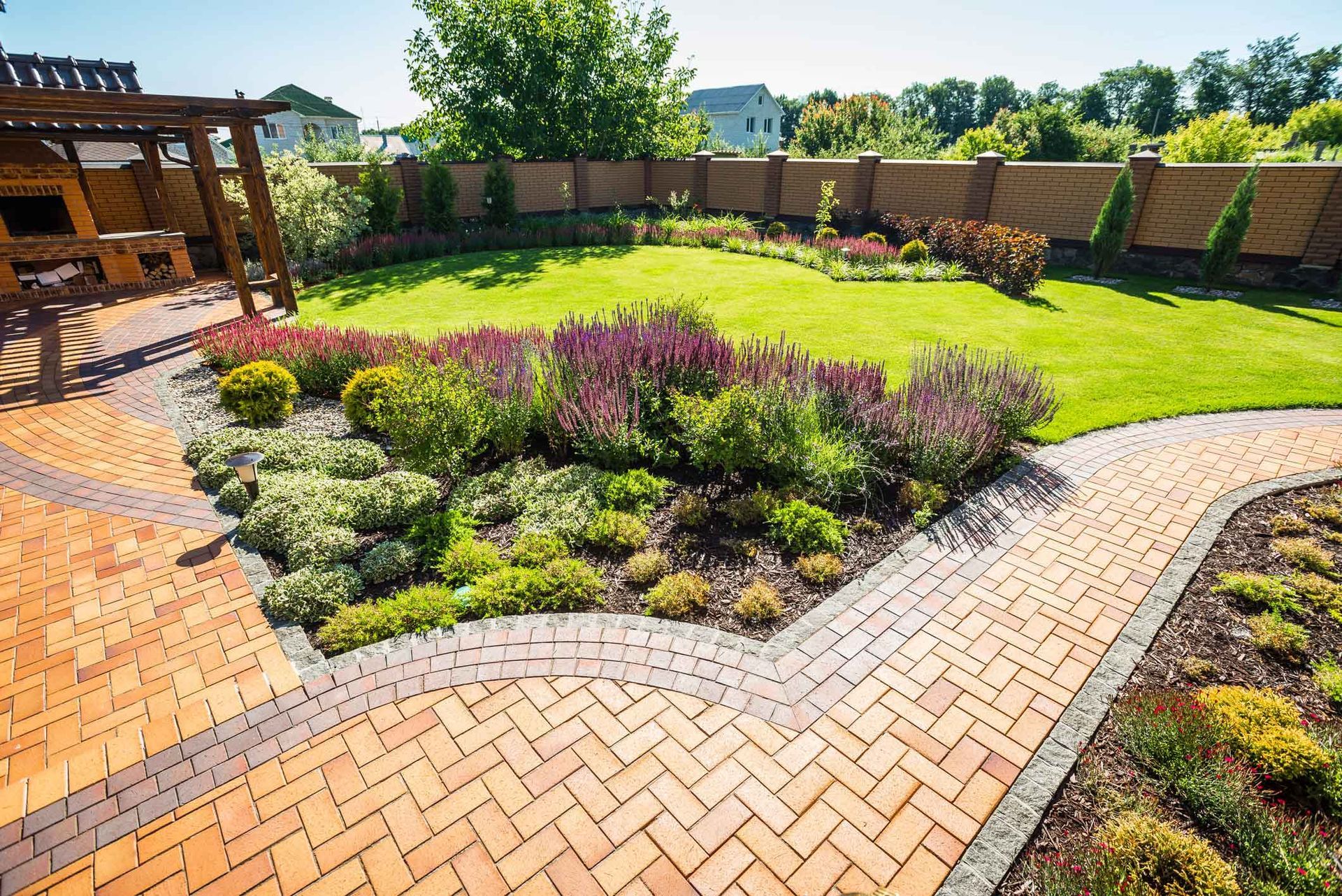 A sunny backyard with a herringbone-patterned brick patio, a curved flower bed with shrubs, and a lush green lawn.
