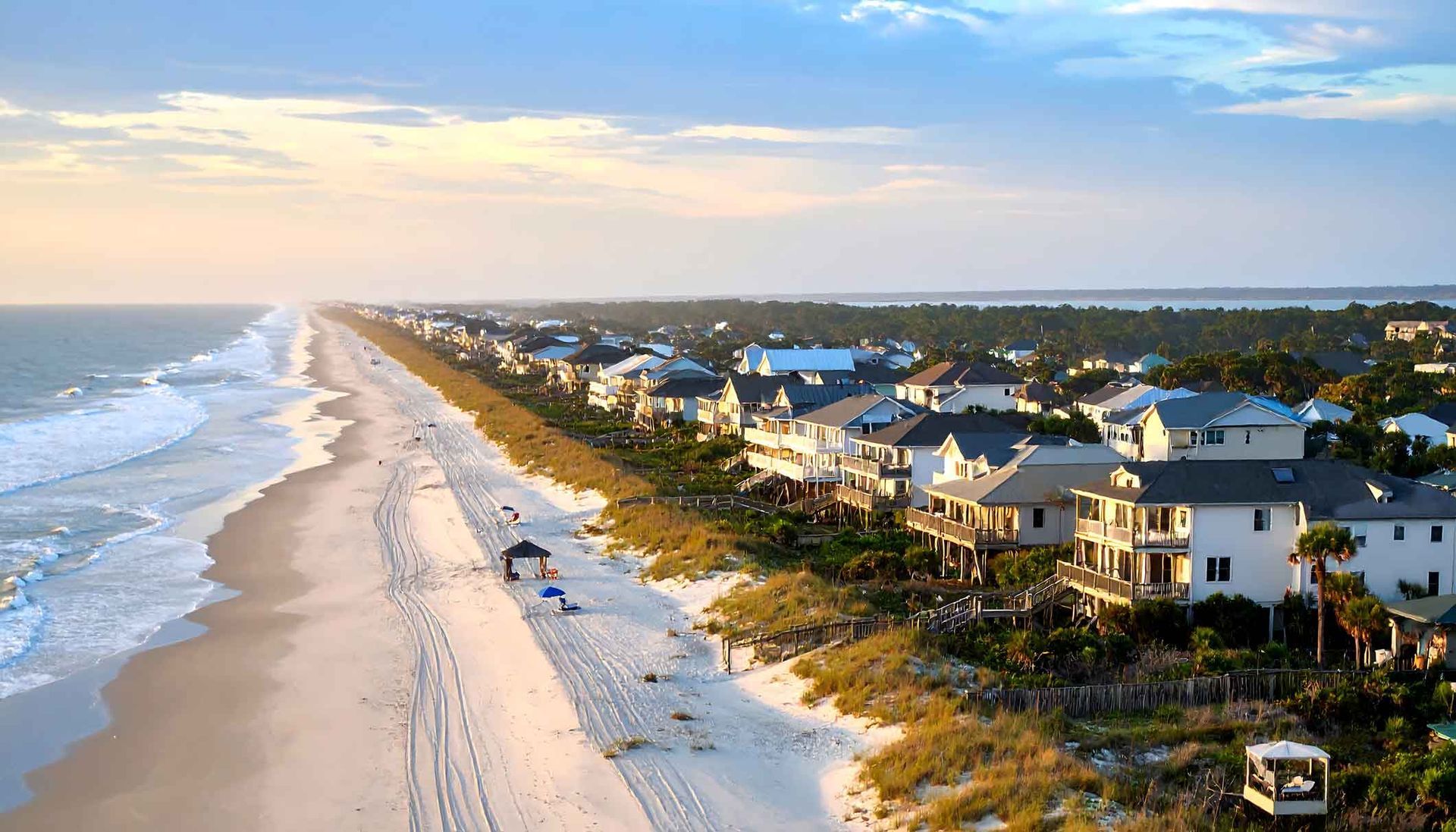 Aerial view of a coastal beach neighborhood with rows of houses nestled along the sandy shoreline at sunset. Aerial view of a coastal beach neighborhood with rows of houses nestled along the sandy shoreline at sunset.