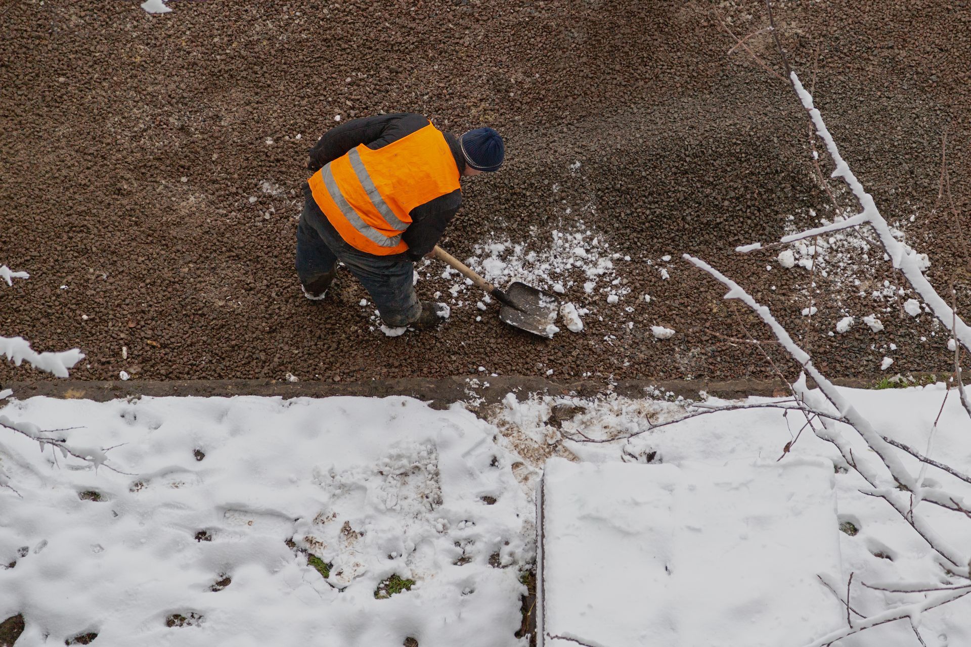 A person in a bright orange high-visibility vest uses a shovel to clear snow from a gravel area on a winter day.