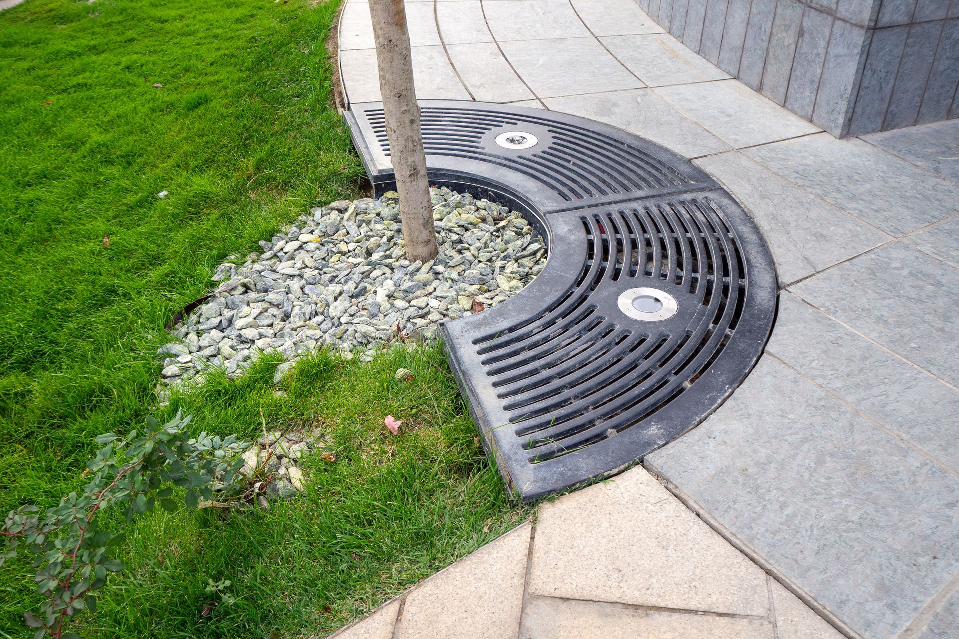 A semi-circular metal tree grate with integrated lights surrounds a tree base, set between stone paving and a grass lawn.