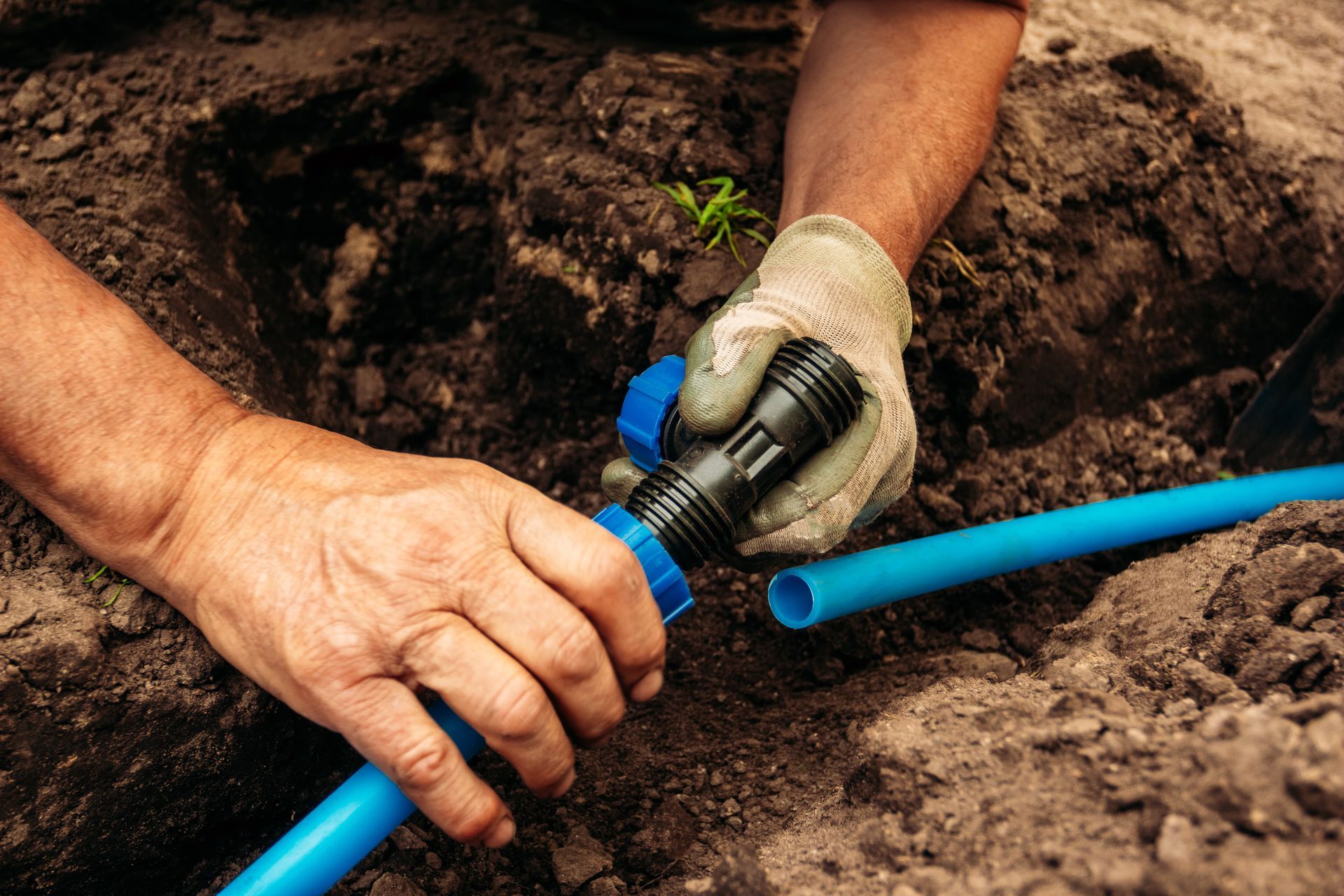Hands in work gloves join blue plastic pipes with a black connector fitting in a trench of soil.