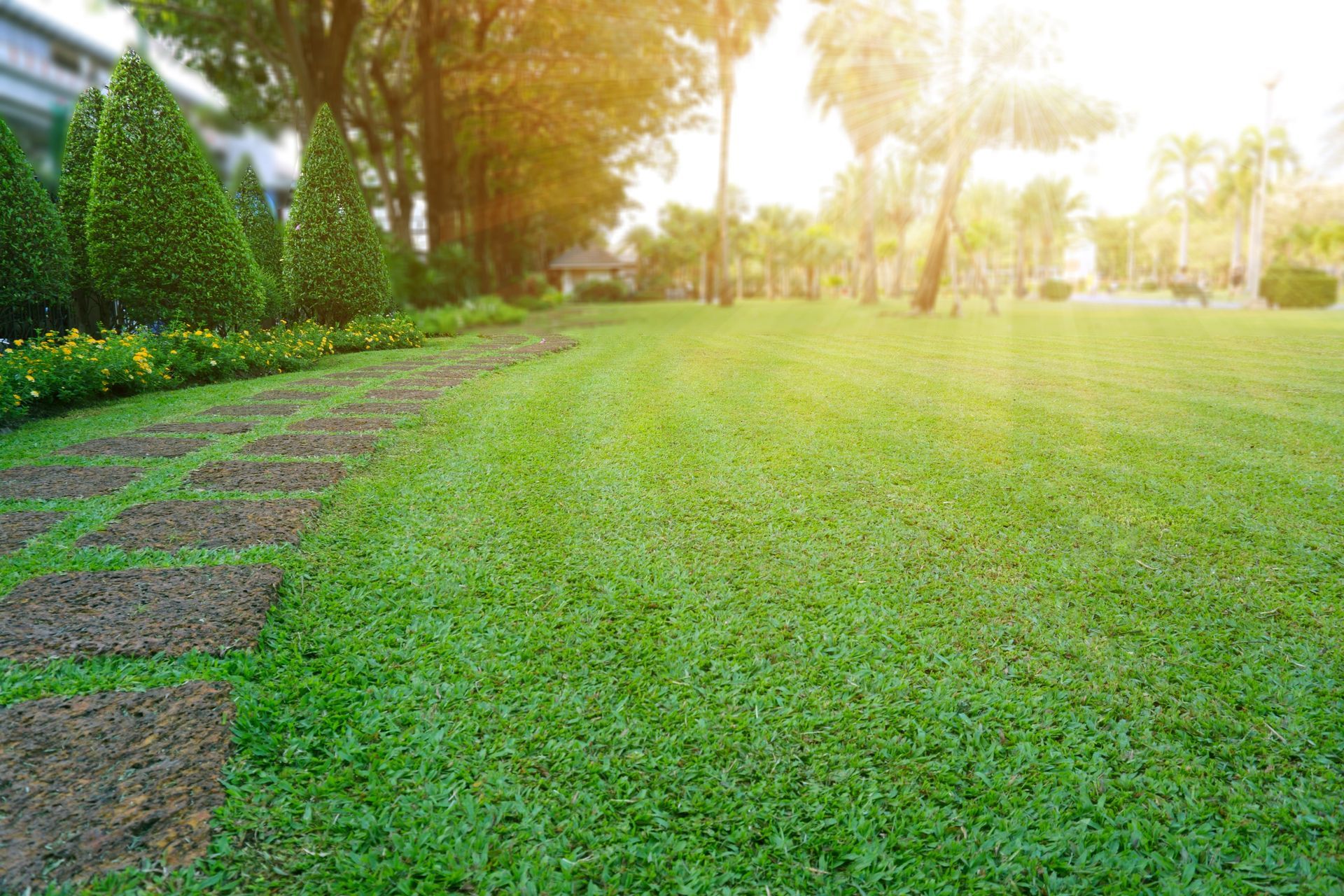A stone path curves through a lush green lawn in a park at sunset, framed by neatly trimmed hedges and trees.