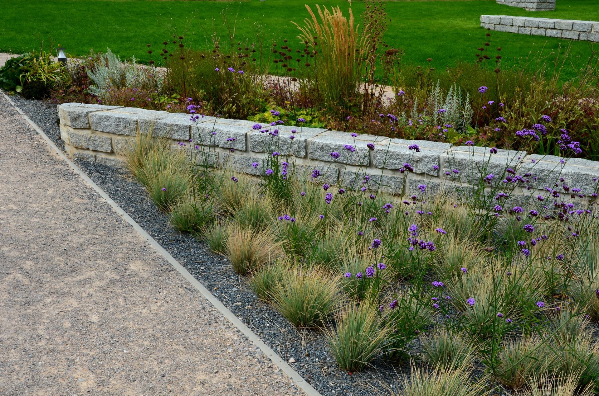 A landscaped garden with a gravel path, a tiered stone retaining wall, and purple wildflowers bordering a green lawn.