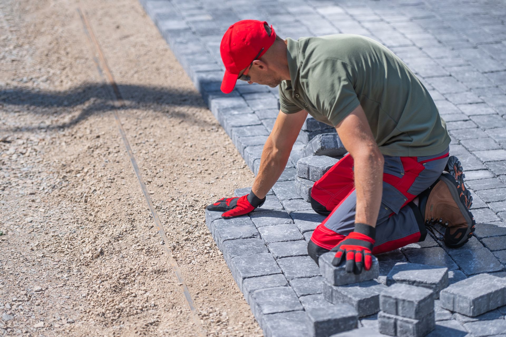 A person in a red cap and work clothes kneels, installing grey paving stones next to a gravel base.