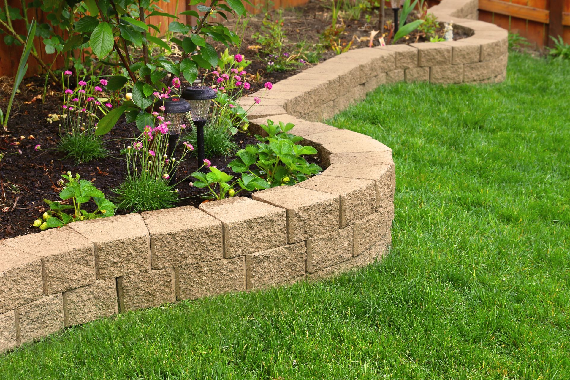 A curved garden retaining wall made of tan, textured stone blocks borders a lush green lawn and flower bed.