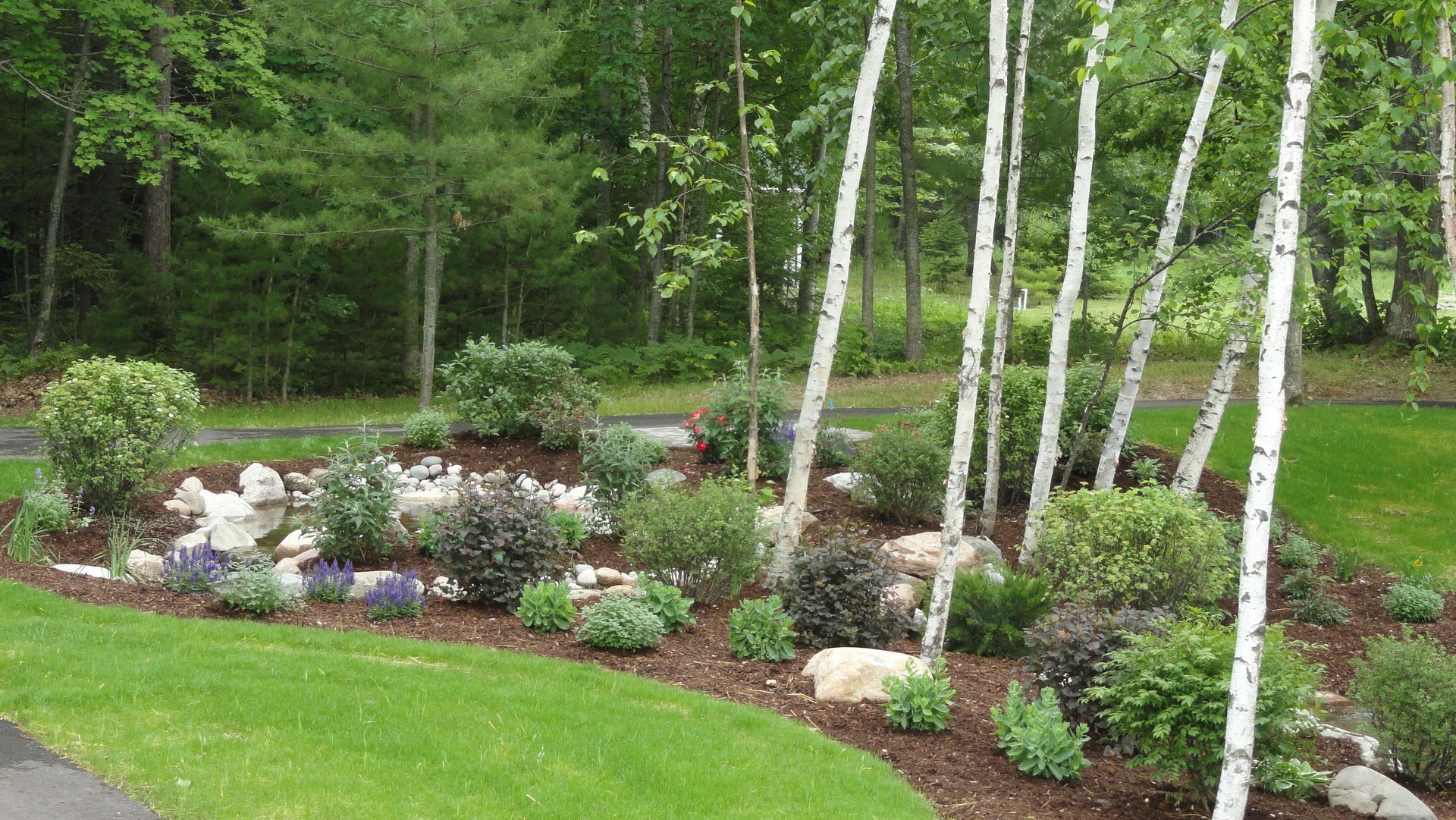 A person in green work pants uses a shovel to lift a pile of dark soil in a garden.