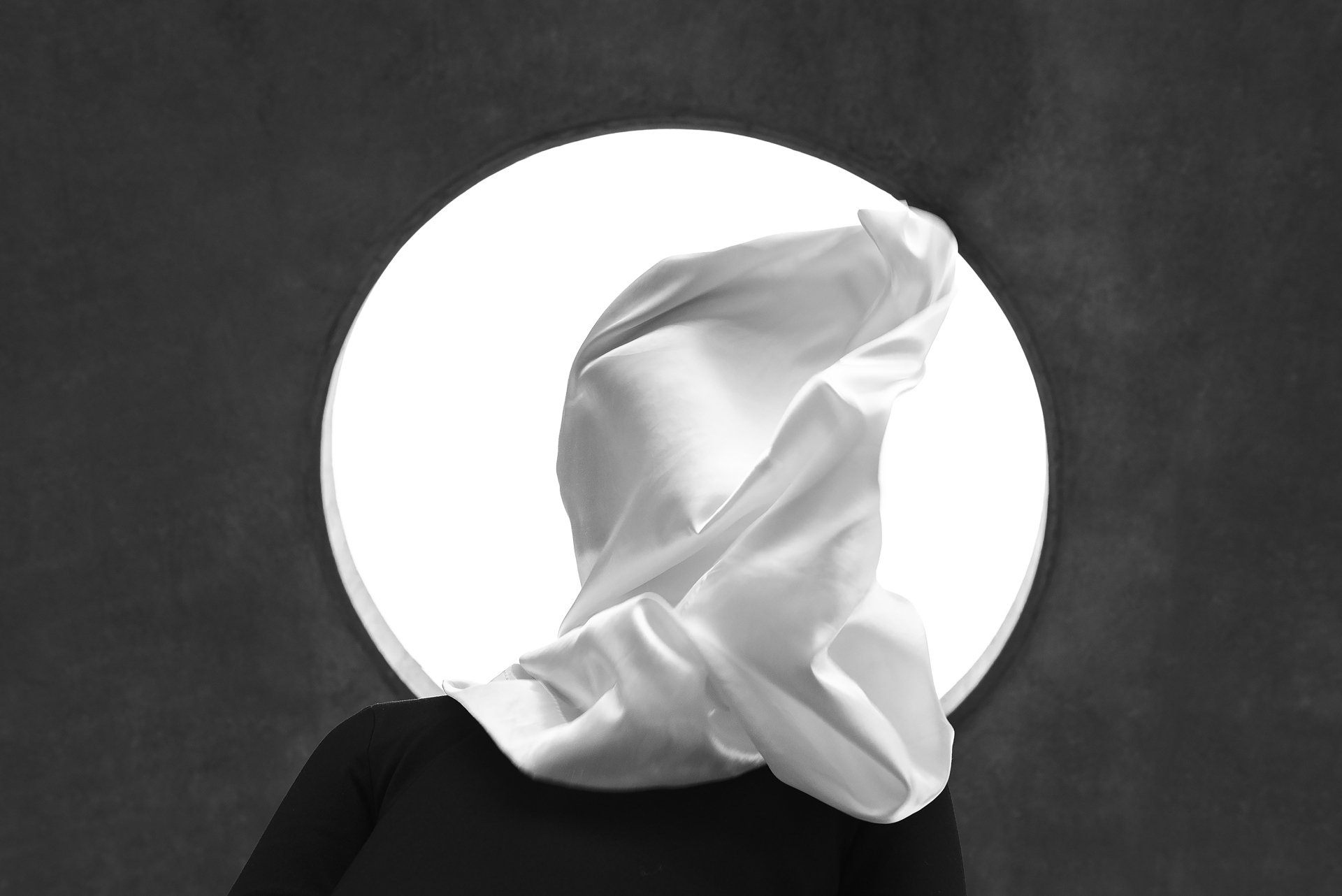 Person wearing a white draped hood in front of a bright circular light.