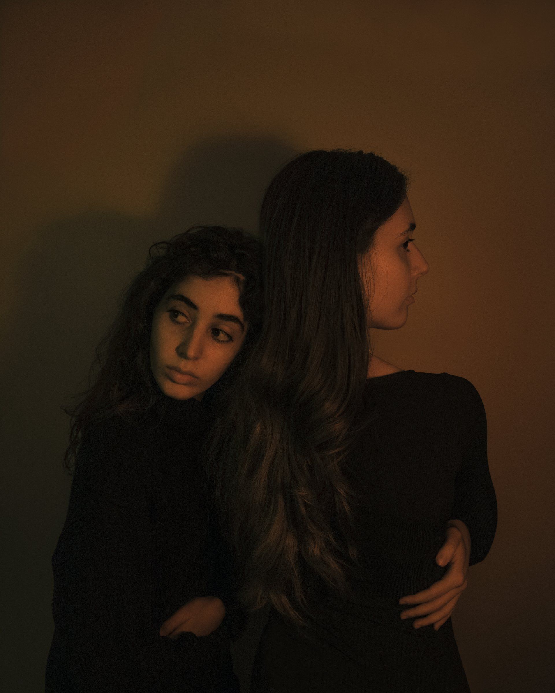 Two people embrace, lit with warm tones. One looks at the camera, the other away, against a neutral background.