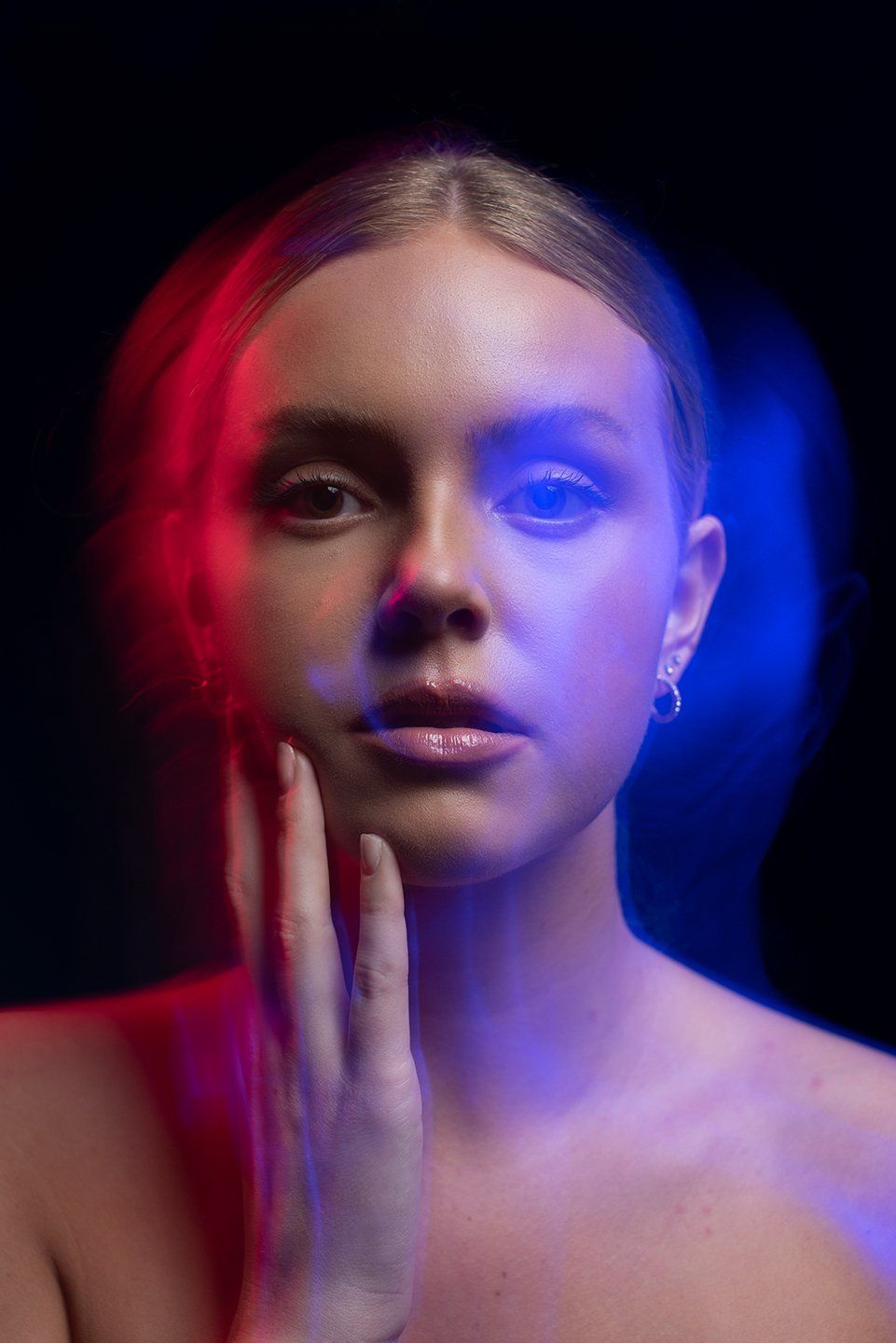 Woman's face with red and blue light streaks. One hand touches her face, black background.