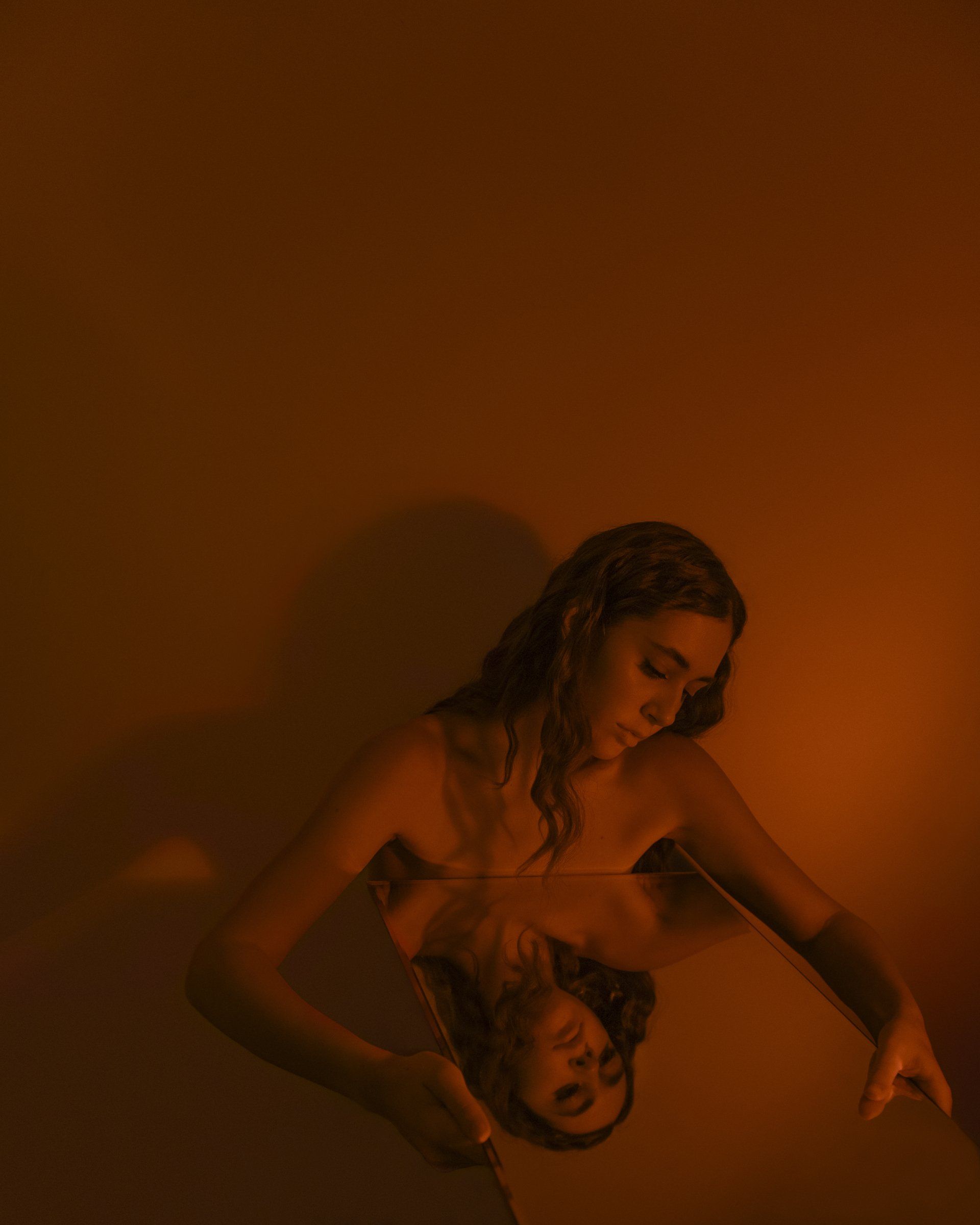 Woman gazing at her reflection in a tilted mirror, lit with warm orange tones.