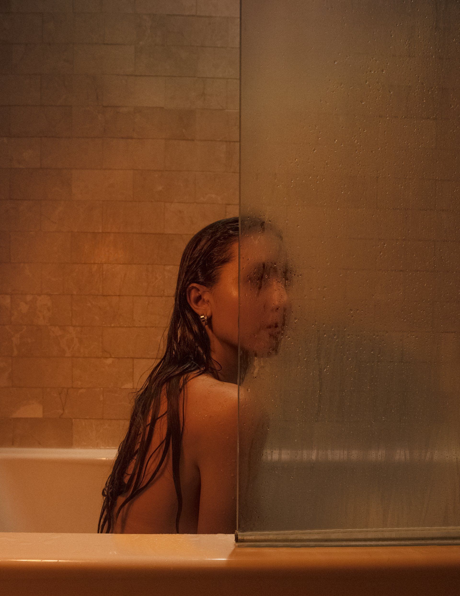 Woman in a steamy shower, leaning against glass, gazing toward the camera.