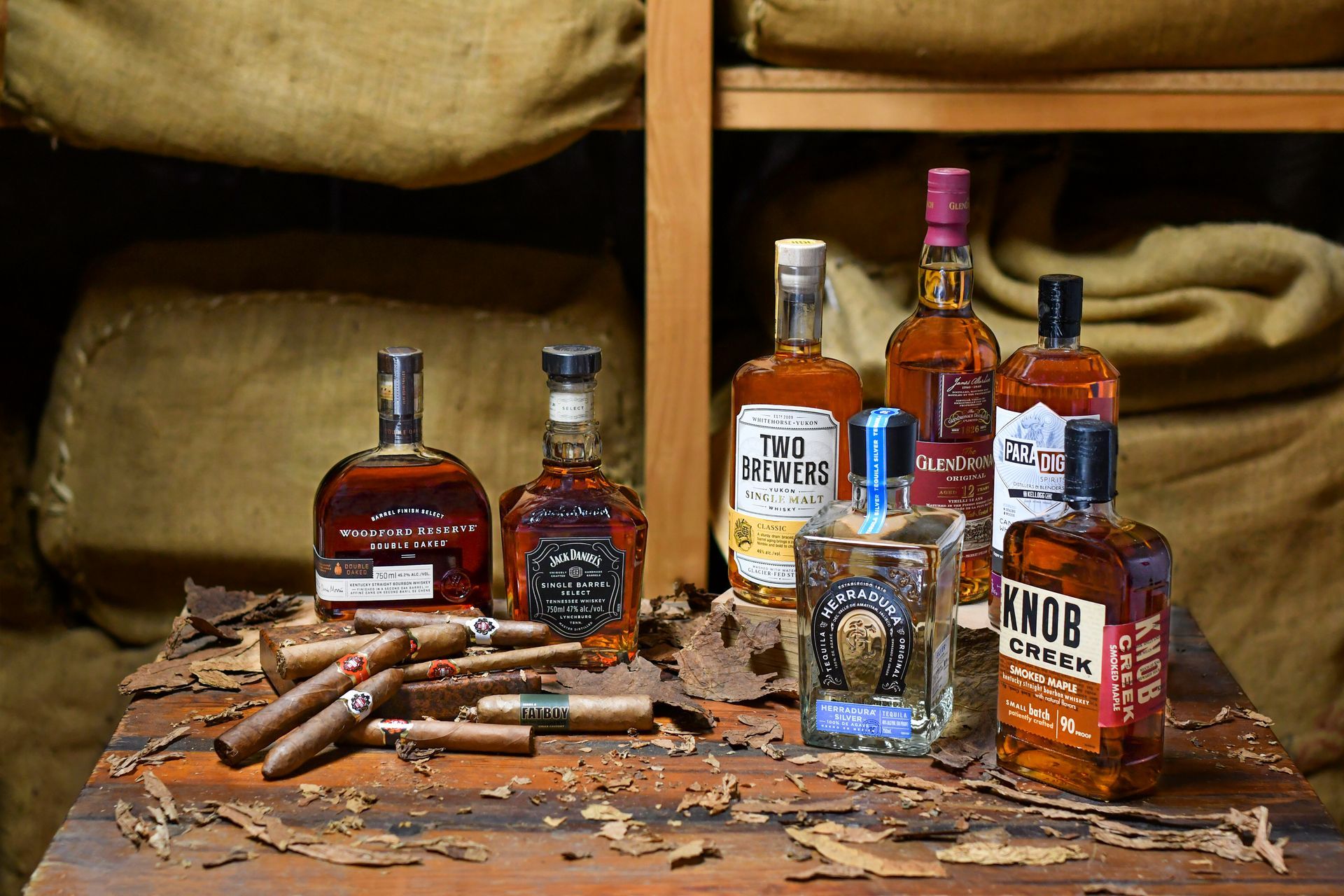 Bottles of bourbon and cigars arranged on a weathered wooden surface, burlap sacks in the background.