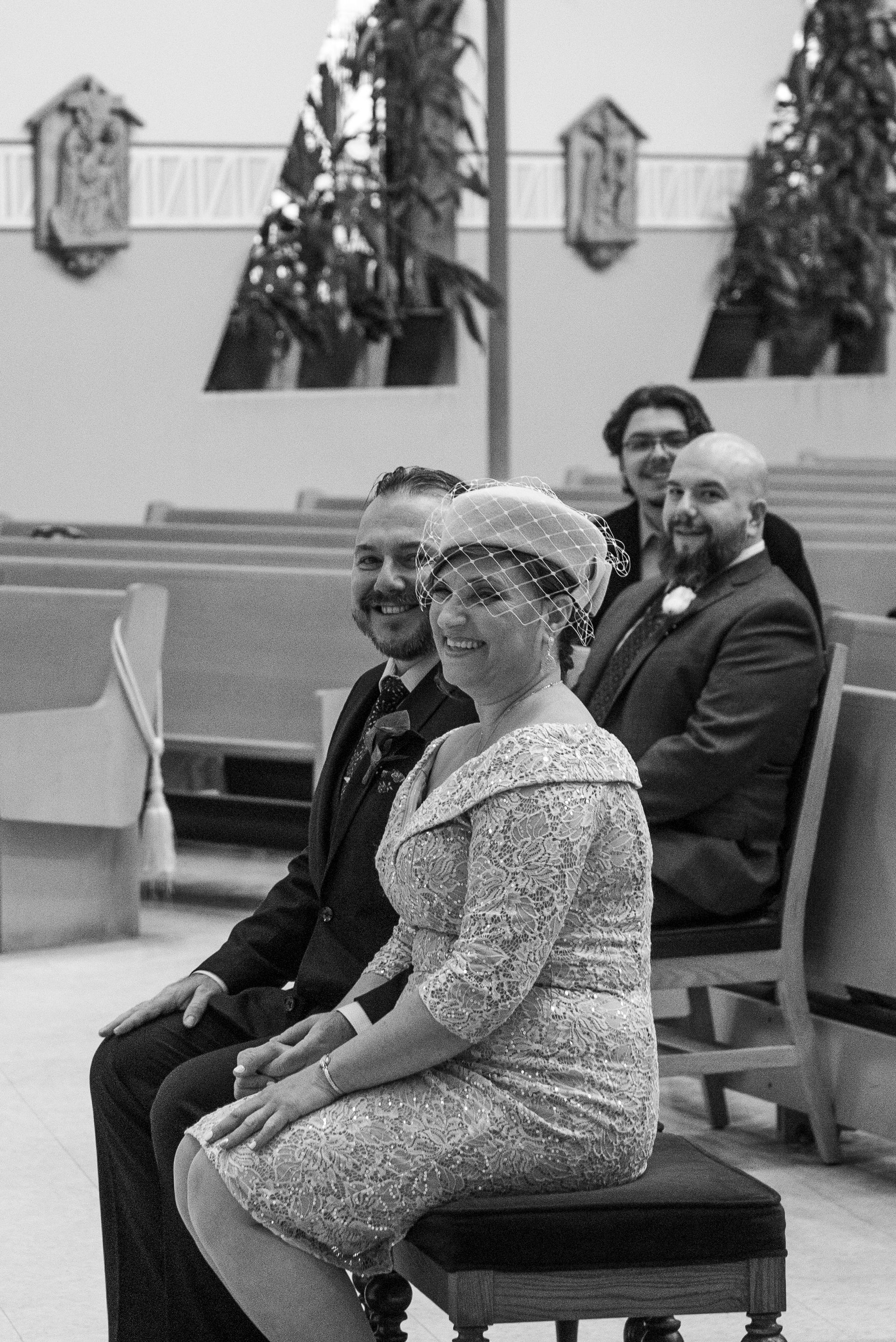 Wedding guests seated in a church. Woman in a dress and hat smiles. Men in suits behind her.