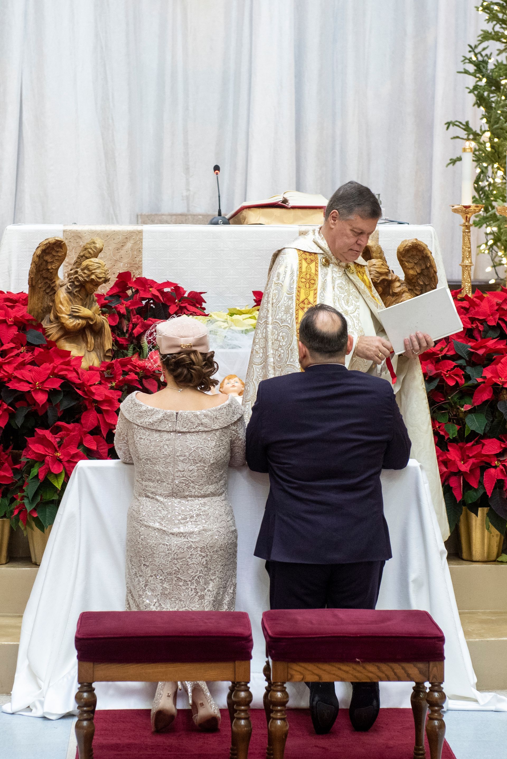 Couple kneels at altar during a church ceremony; priest in vestments holds a document.