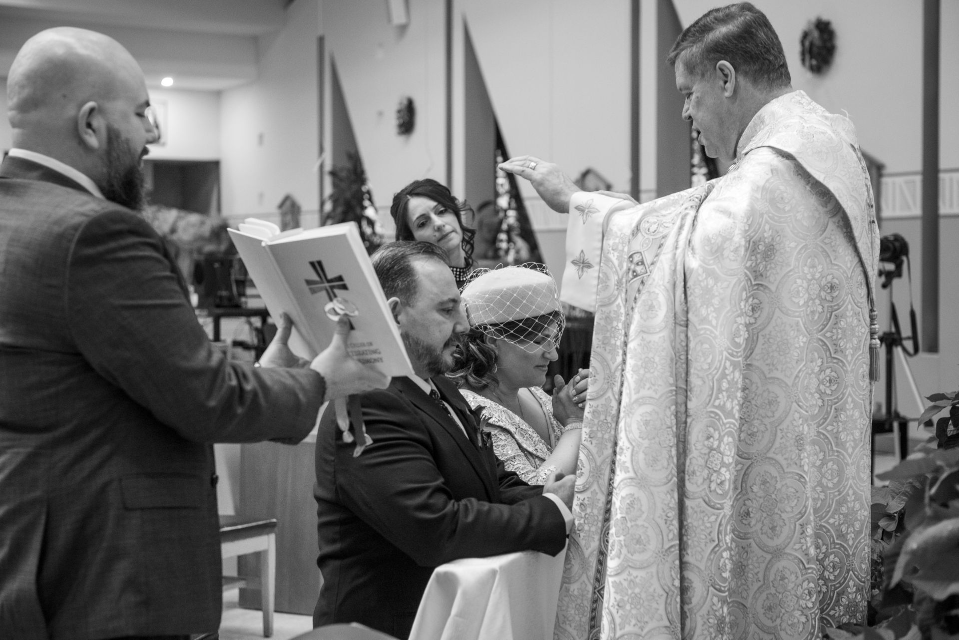 Wedding ceremony: Priest blesses couple with assistant holding book, other guests watch.