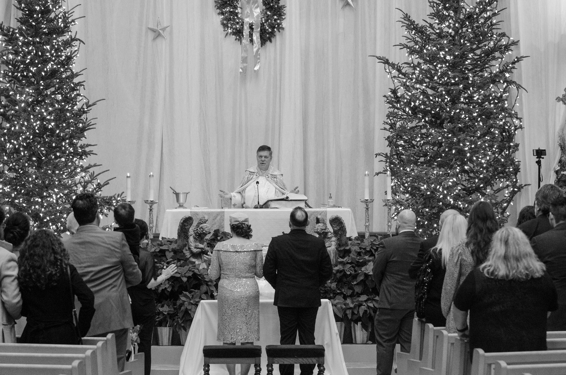 Wedding ceremony in a decorated church; couple at altar, officiant, guests, and Christmas trees.