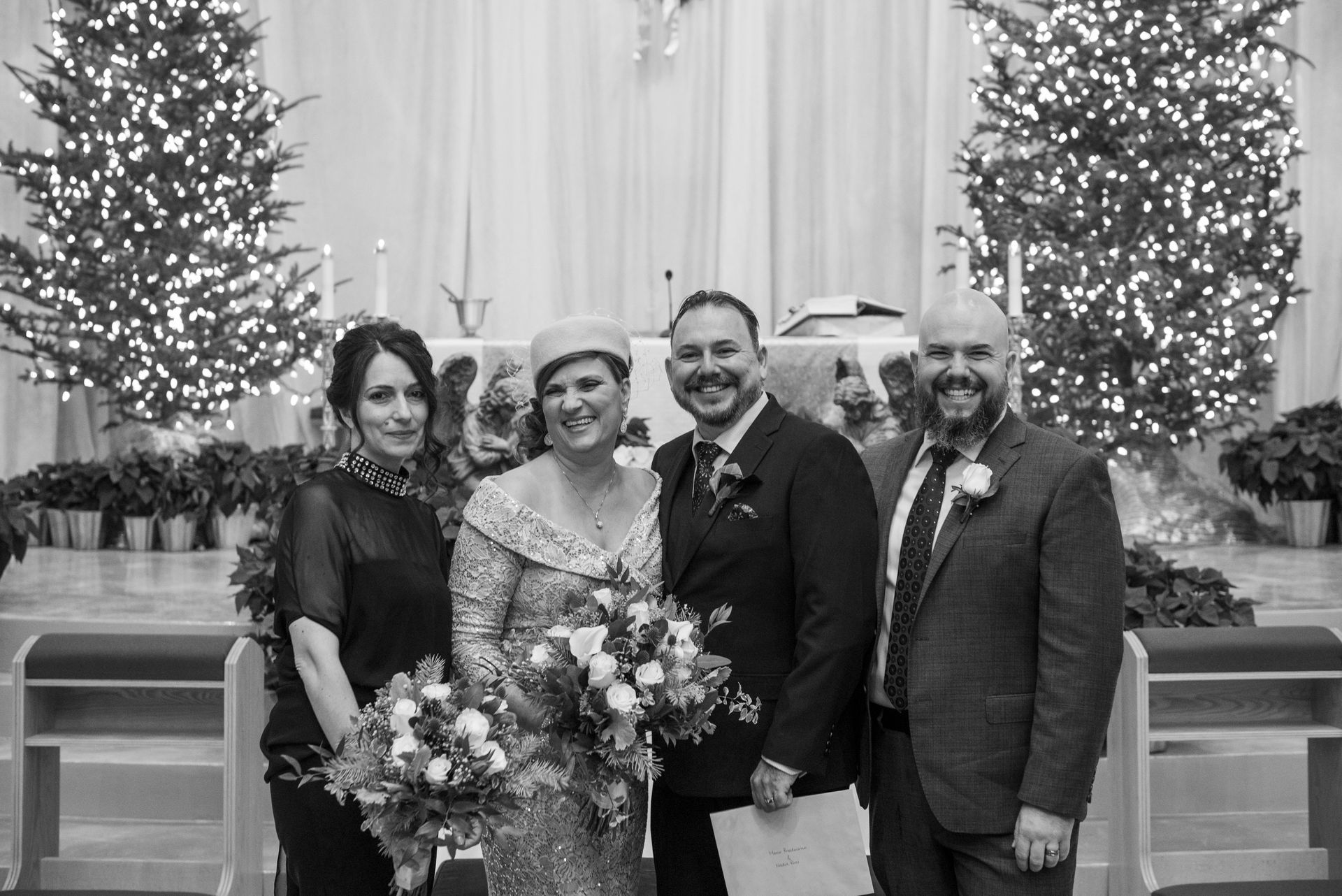 Four people pose in a church: two brides, a groom, and another person with Christmas trees in the background.