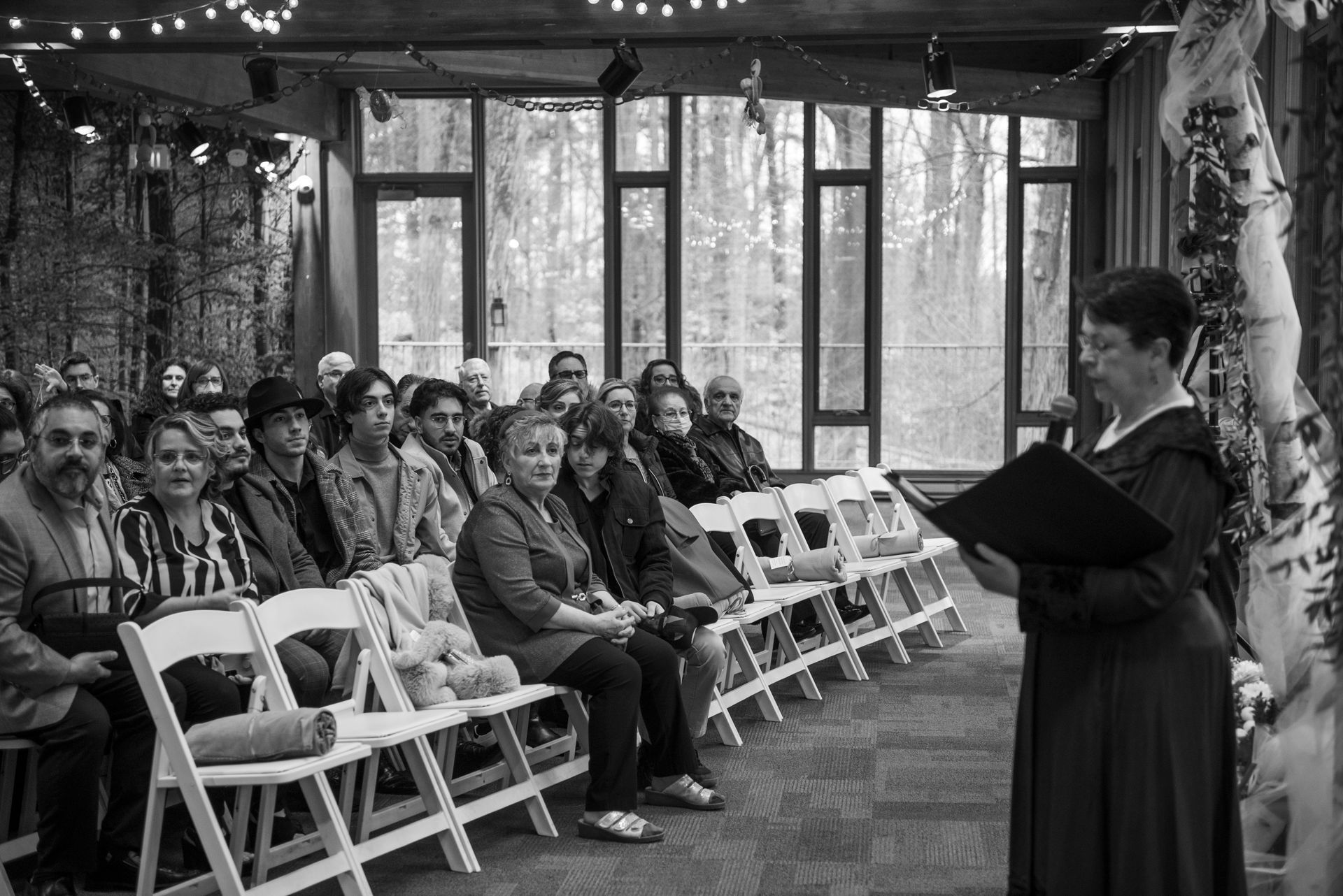 A person reads from a book, addressing a seated audience in a bright indoor setting.