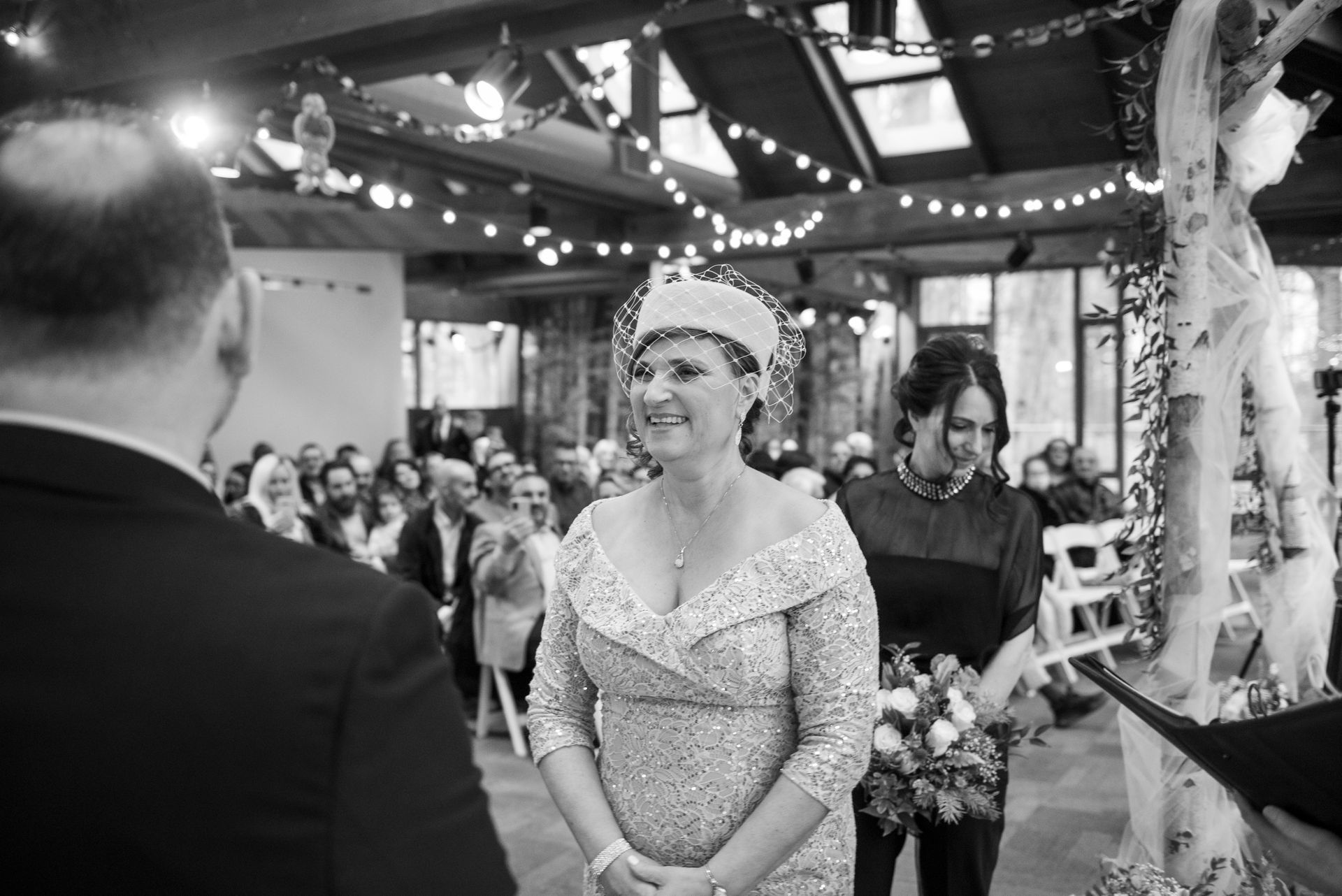 Bride at wedding ceremony, smiles. Wearing beaded dress, hat. Indoor setting with guests and decorations.
