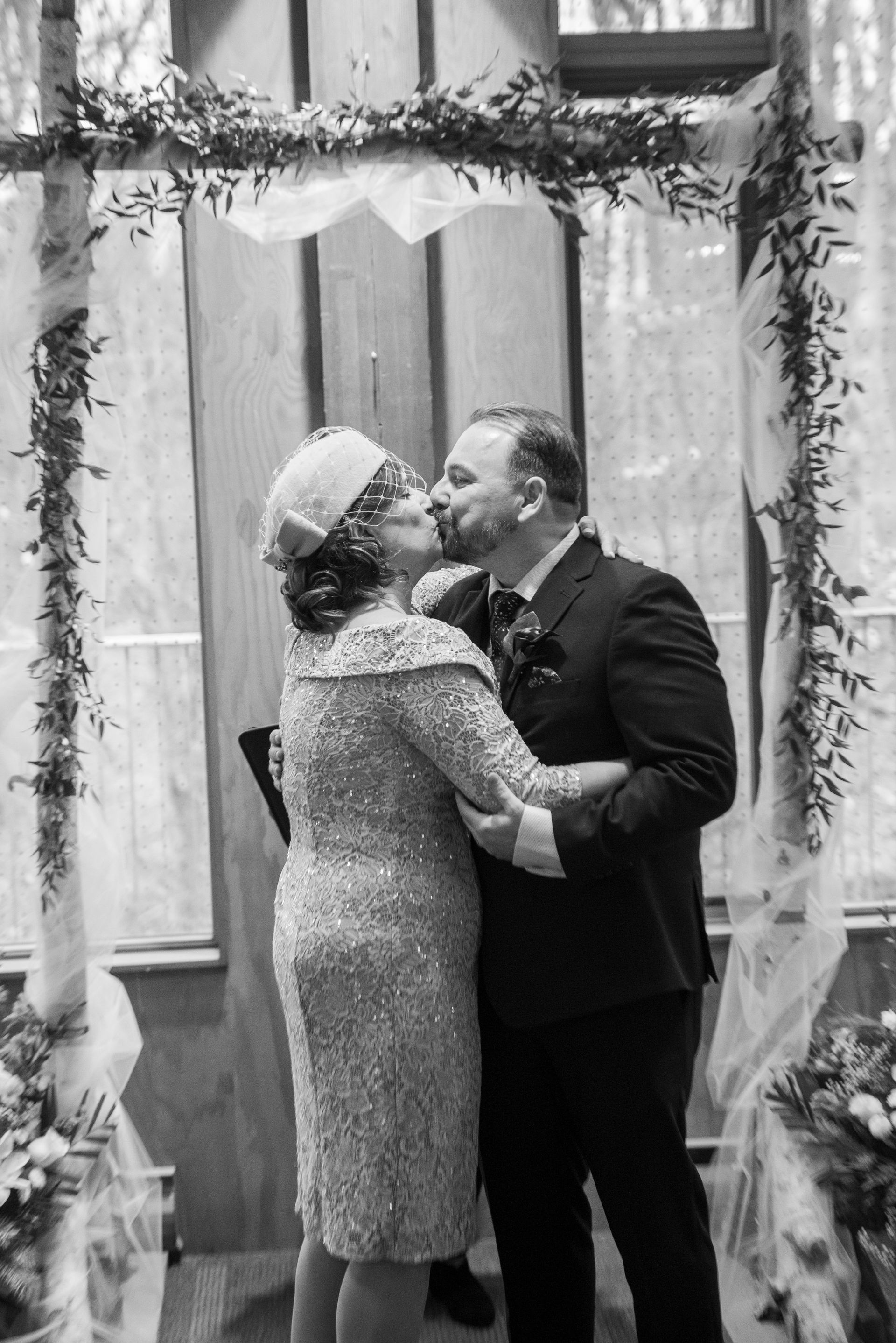 Couple kissing under an archway decorated with greenery; wedding ceremony. Black and white.