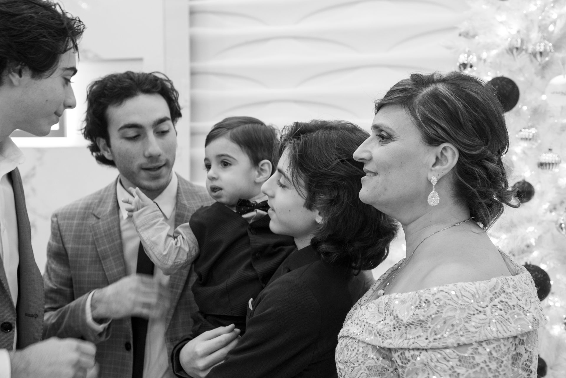 Family gathered, talking near a decorated tree. A person holds a child. Formal attire, black and white.