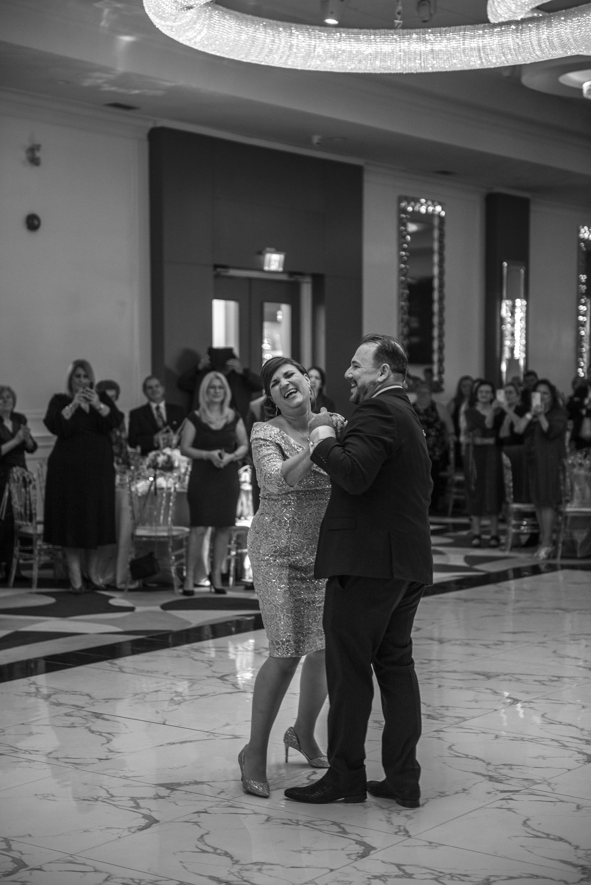 Couple dancing at a reception, smiling, inside a ballroom. Guests are watching and clapping.