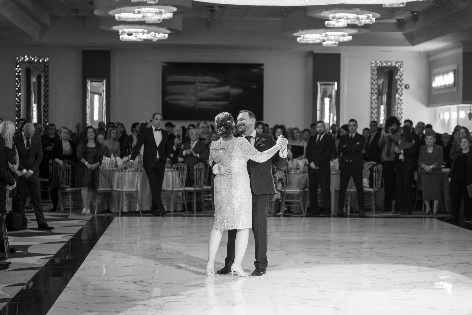 Couple dances on a white floor during a formal event, surrounded by guests in a large ballroom.