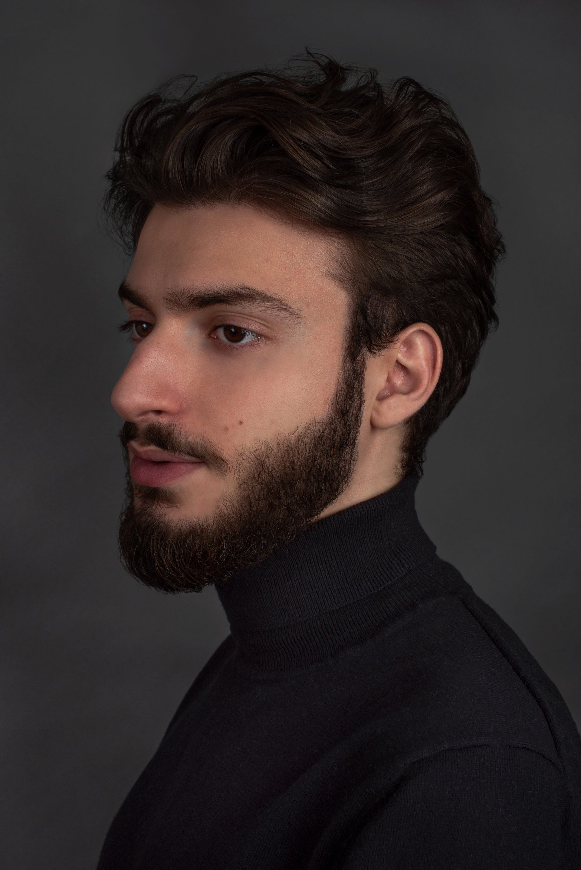 Man with dark wavy hair and beard wearing a black turtleneck, looking to the side against a gray backdrop.