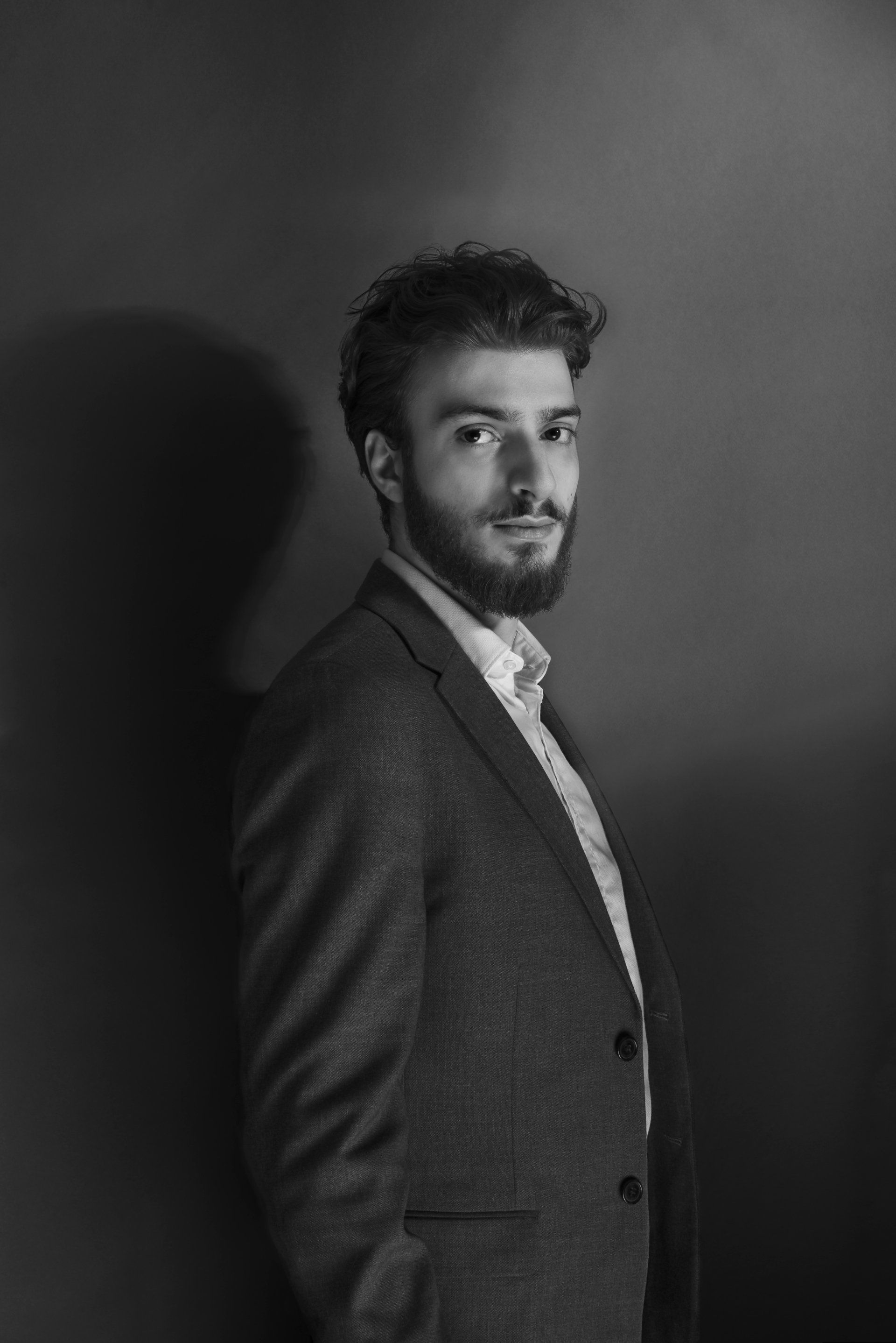 Man in a blazer and open-collared shirt leaning against a wall, looking towards the camera.