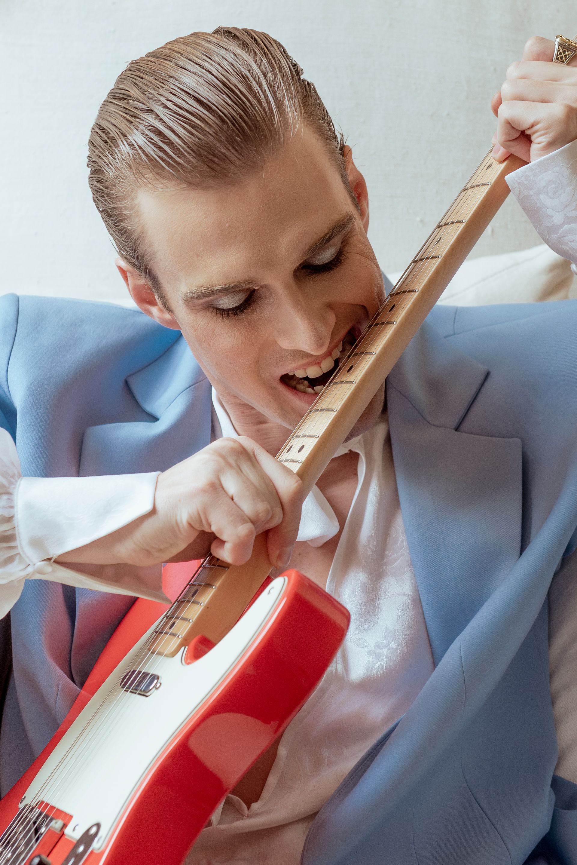 Person in blue suit biting the neck of a red electric guitar, wearing eye makeup.
