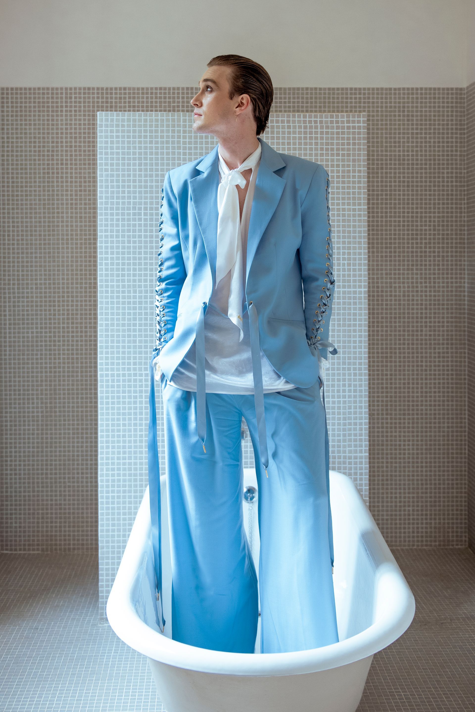 Person in a light blue suit stands in a bathtub, looking upward. White shirt, open jacket, tiled wall.