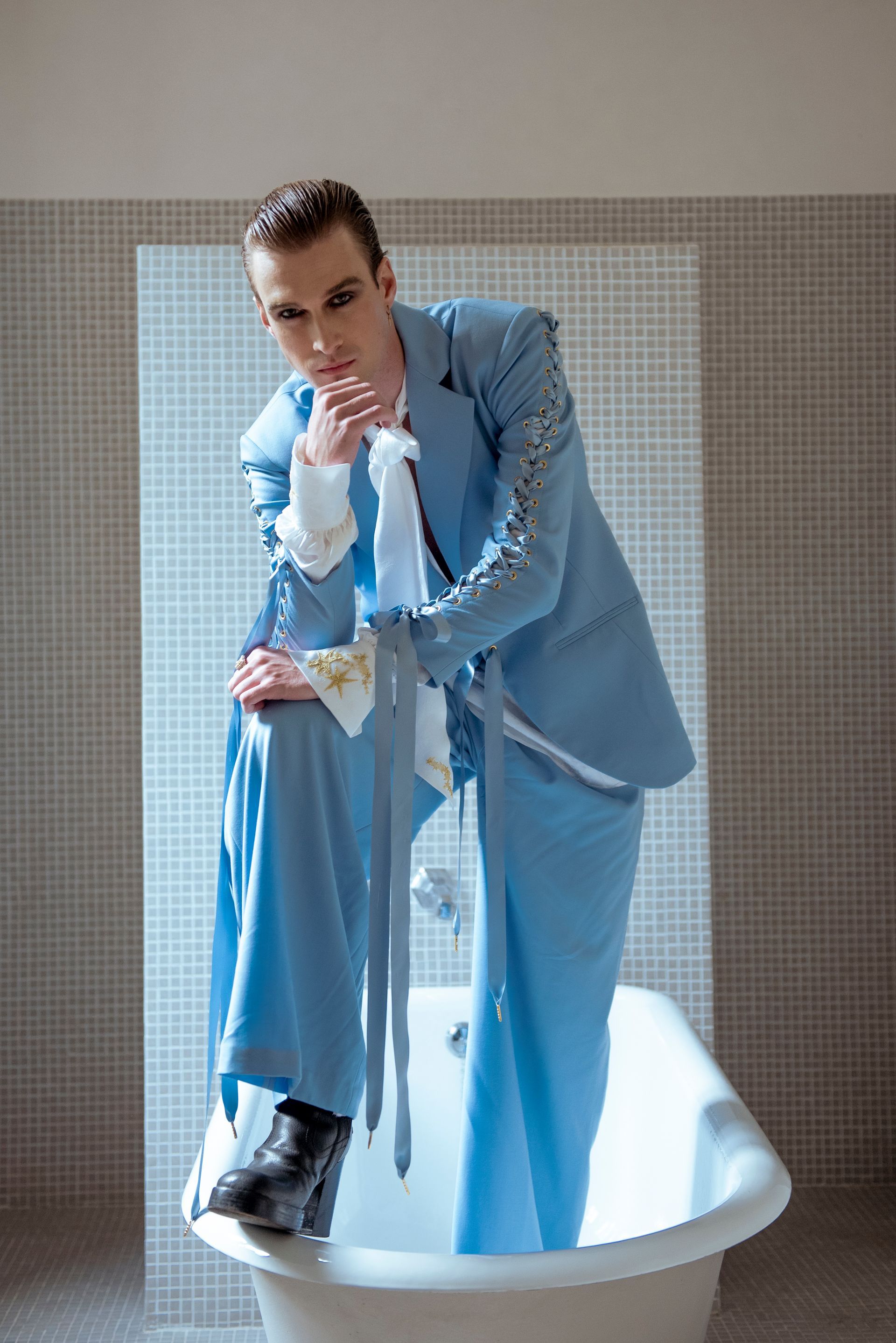 Person in a light blue suit and black boots leans in a bathtub, looking forward.