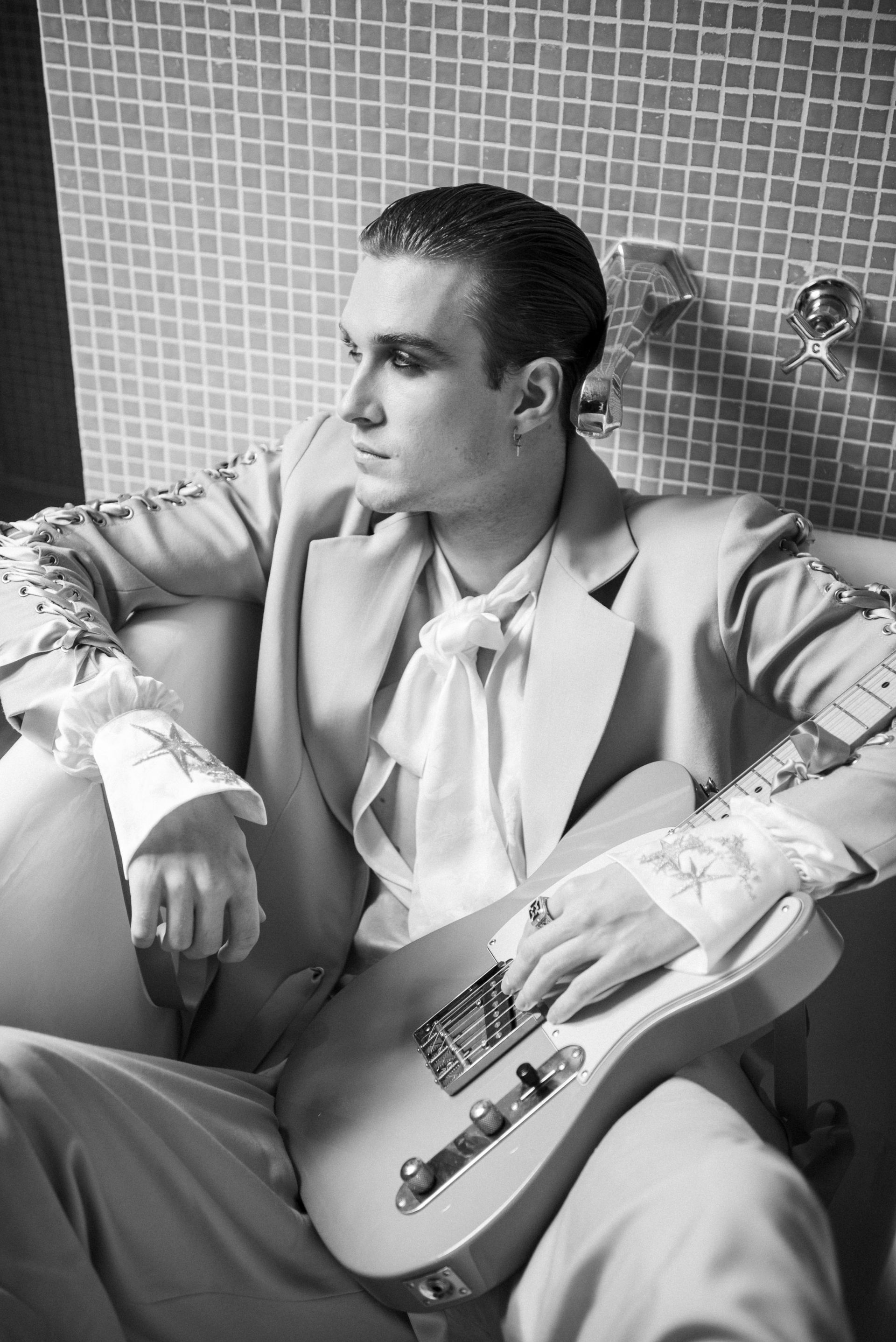 Man in a suit sits in a bathtub, holding a guitar, looking off to the side, black and white.