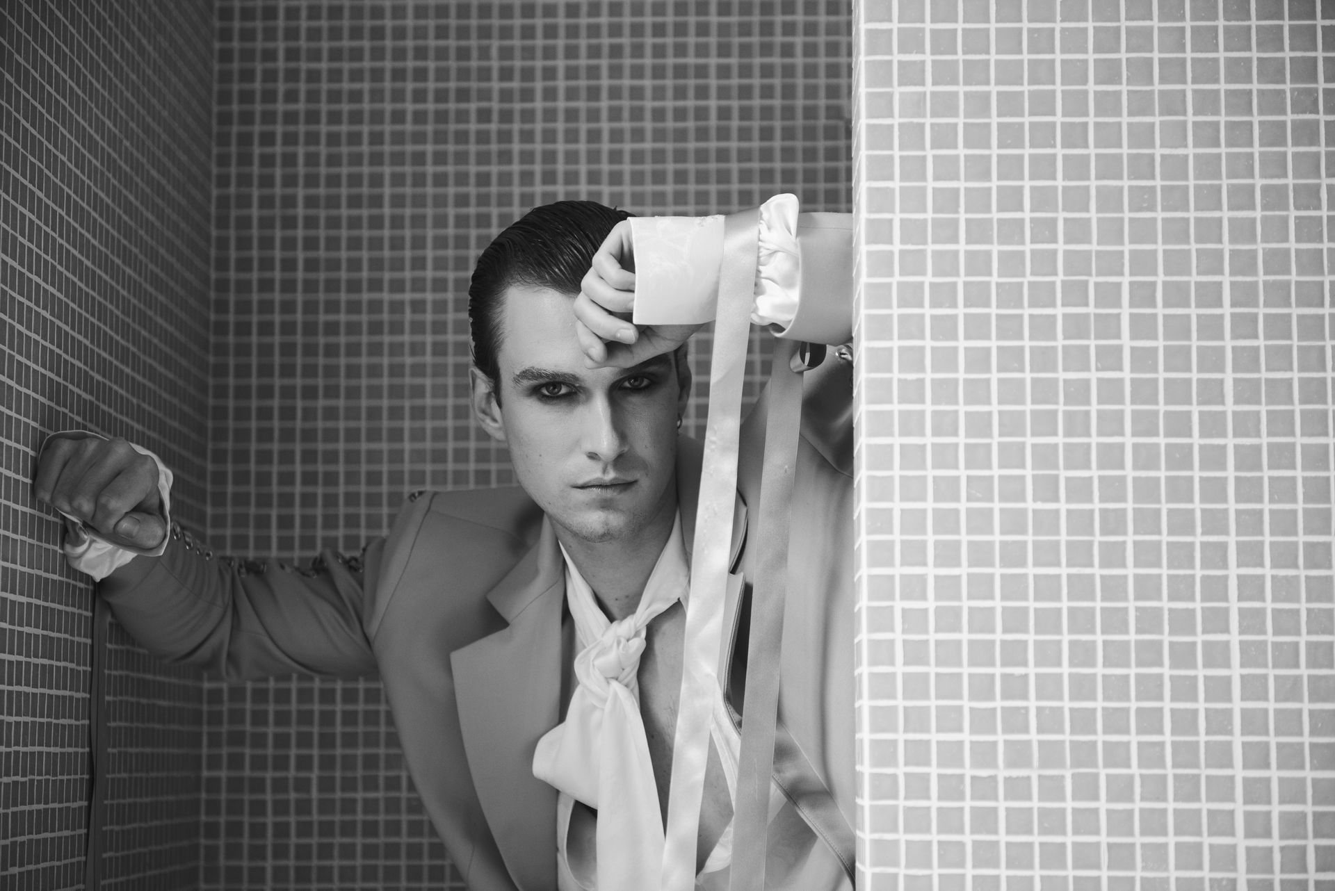 Man in formal wear, dark makeup, leans against tiled wall, holding fabric. Black and white.