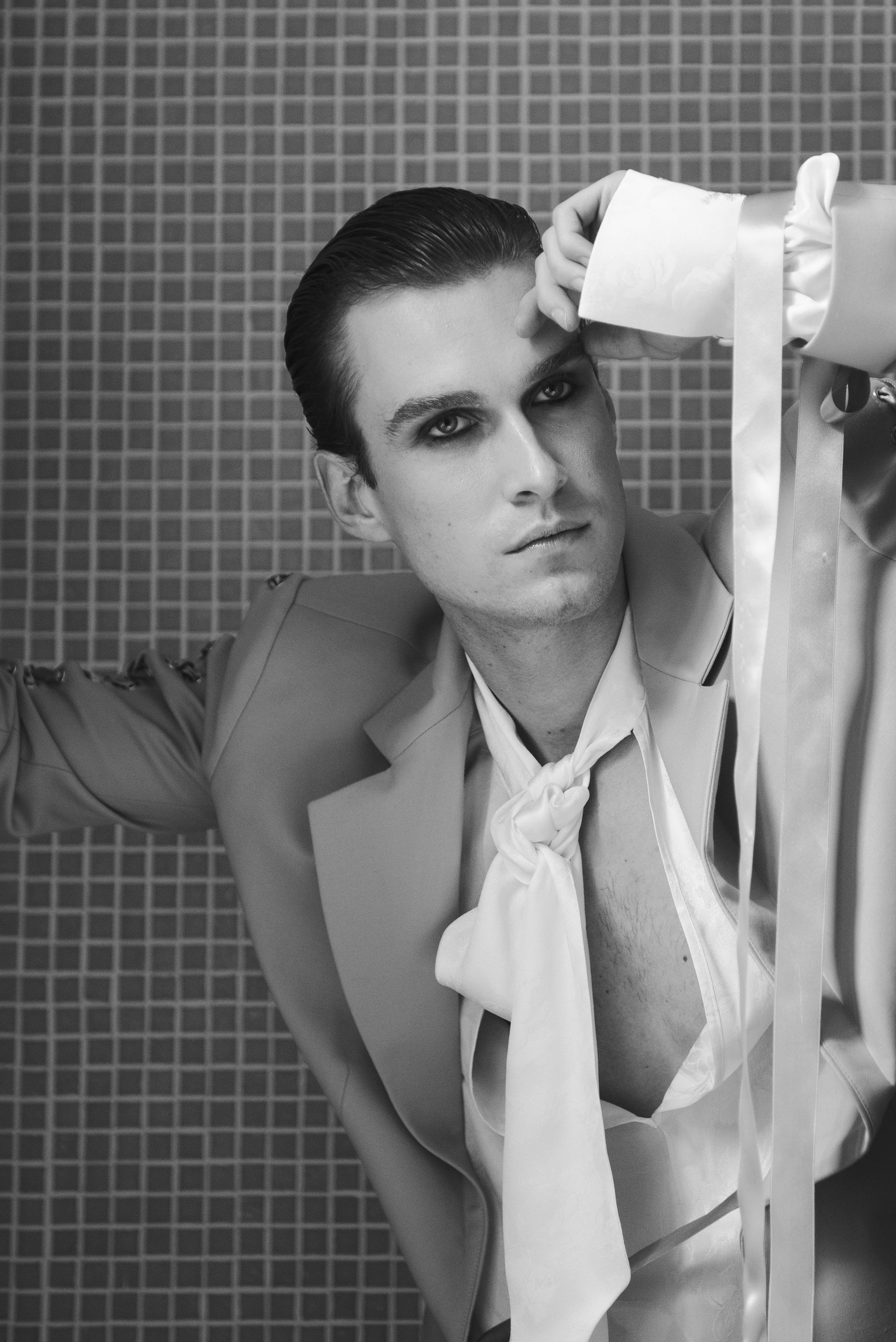 Man in formal attire with dark eye makeup, leaning against a checkered wall, looking away.