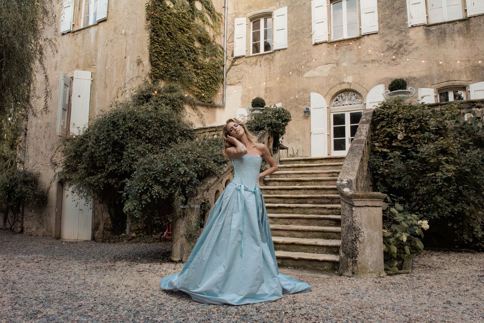 Woman in blue gown stands on stone steps in front of a chateau.