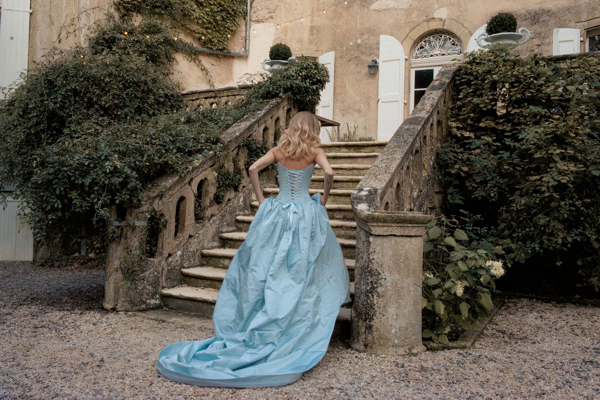 Woman in blue gown climbs stone stairs towards a building.