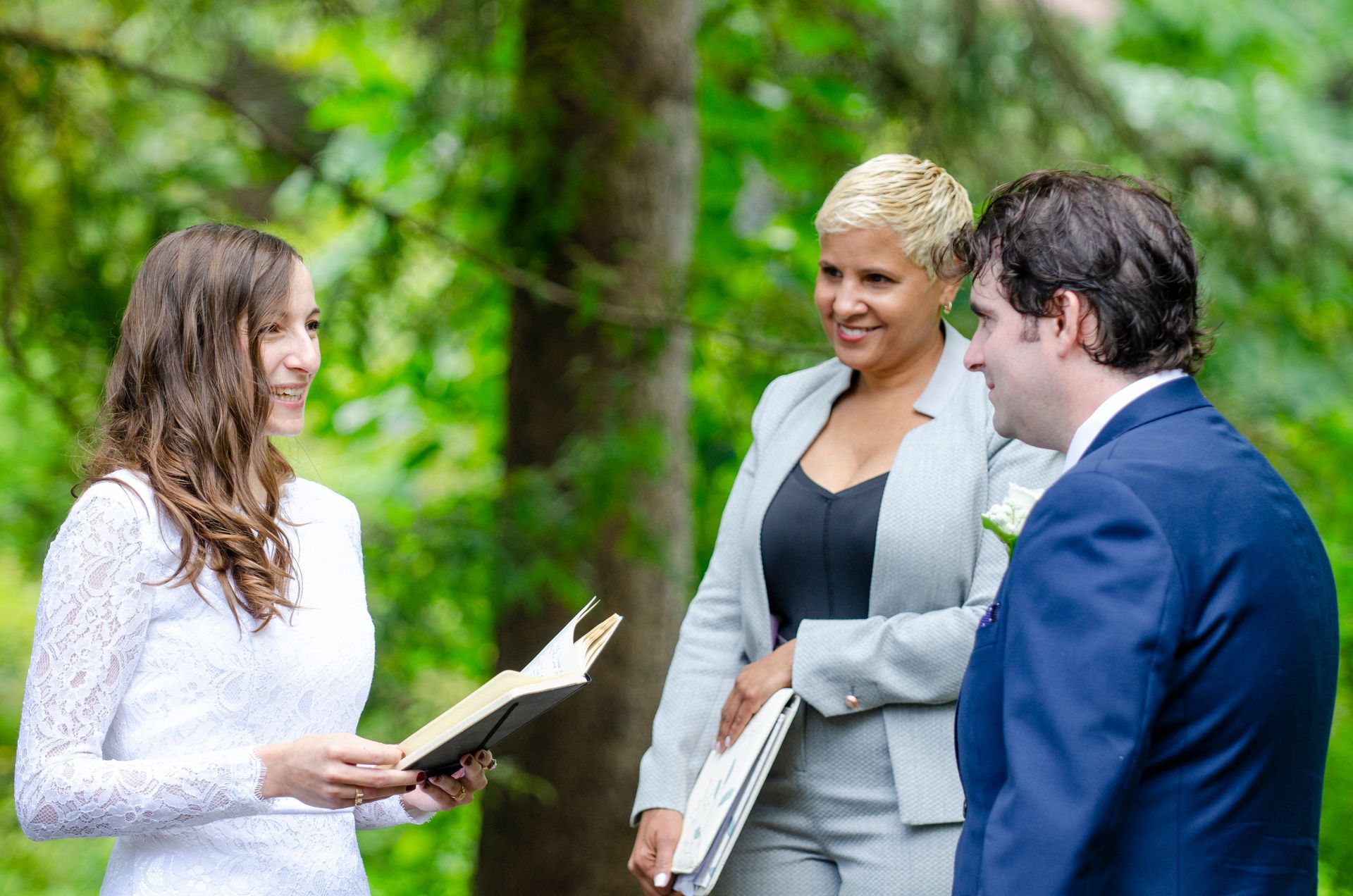 Wedding ceremony with a bride and groom, a celebrant reading vows in a green outdoor setting.