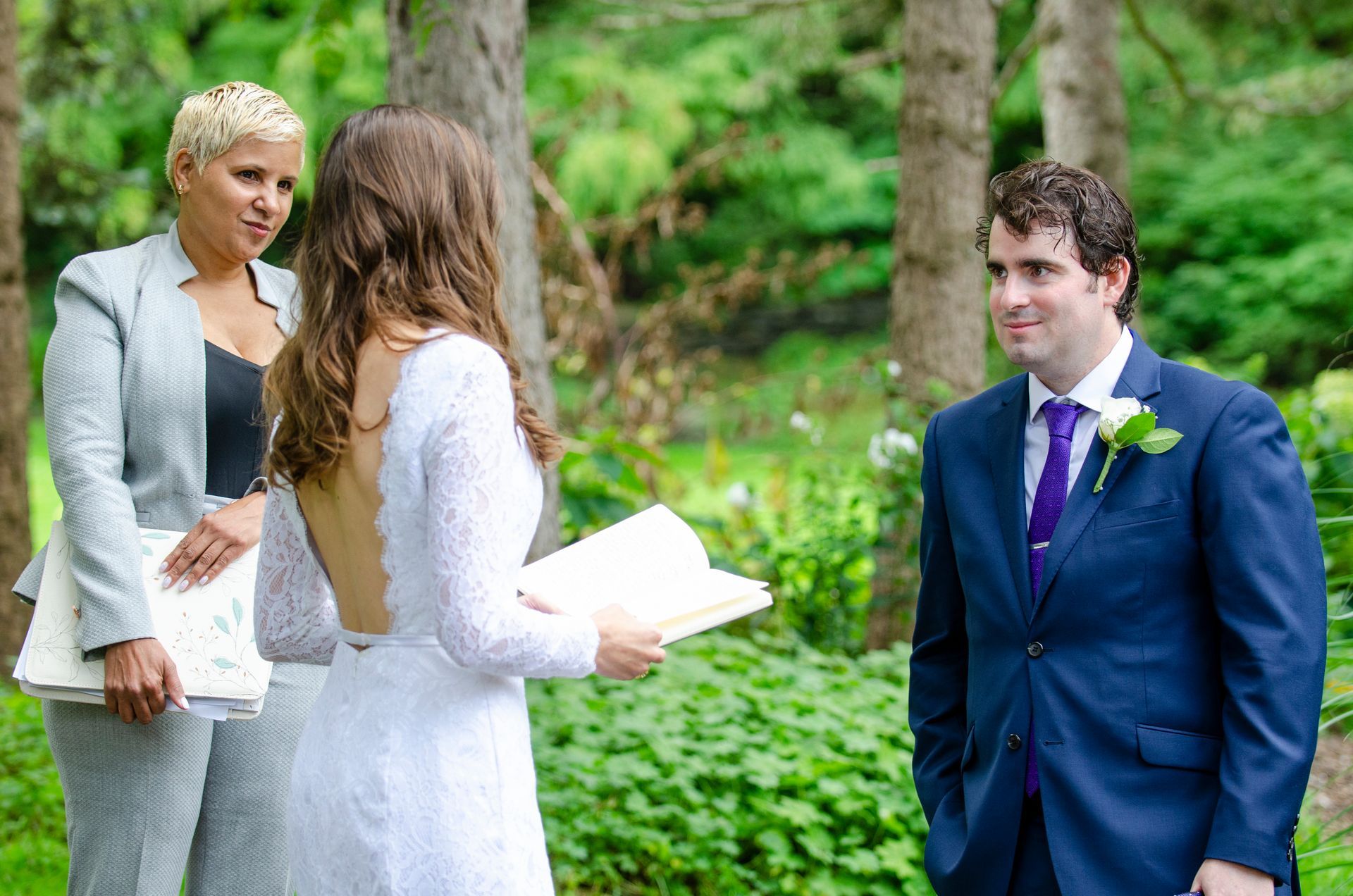 Bride and groom at outdoor wedding ceremony. Bride in white dress, groom in blue suit, officiant in gray.