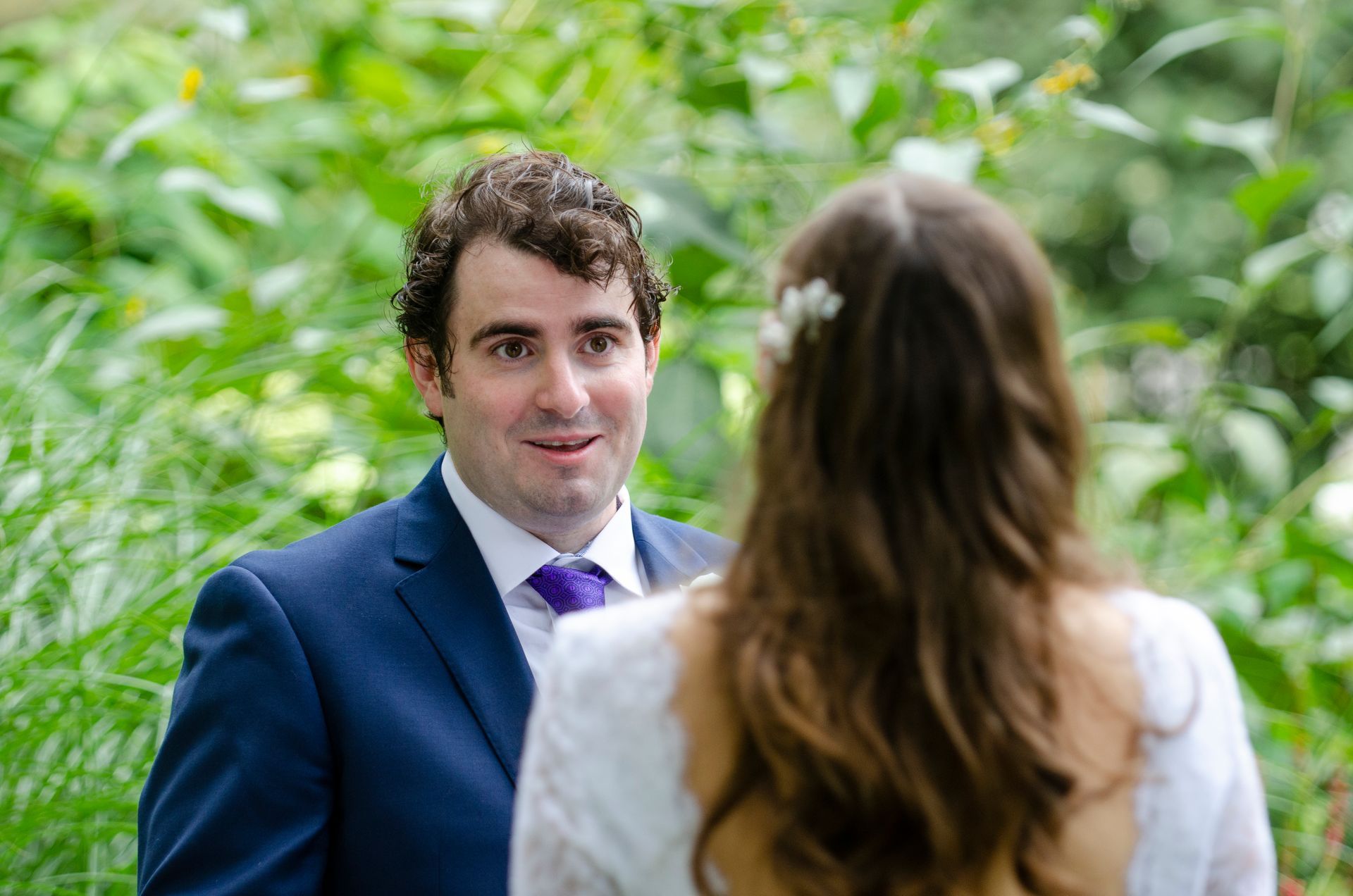 Man in blue suit looks at bride, standing in a garden setting.