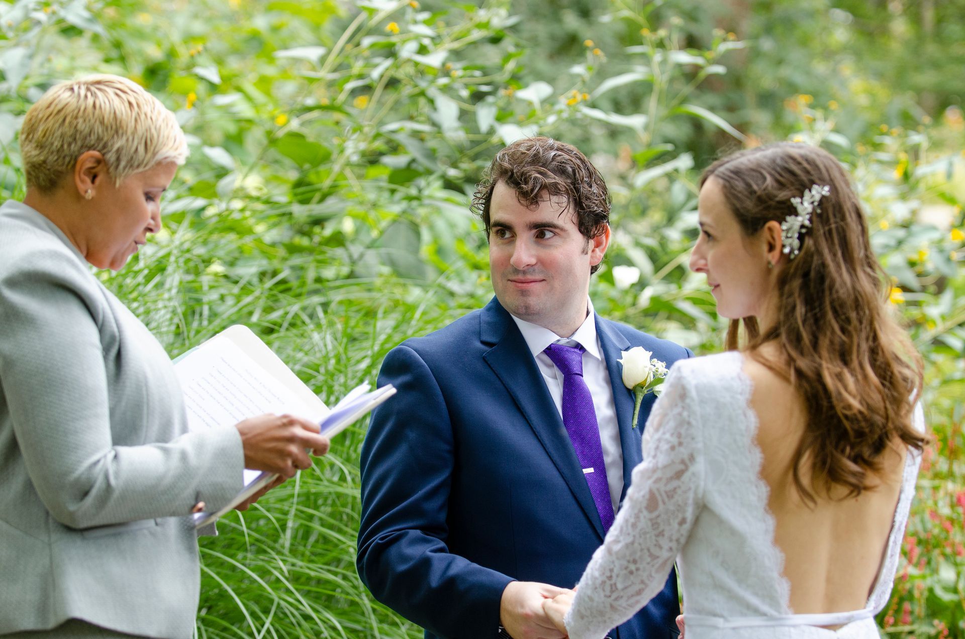 Wedding ceremony outdoors. Couple holding hands, looking at officiant reading from a book. Green foliage in background.