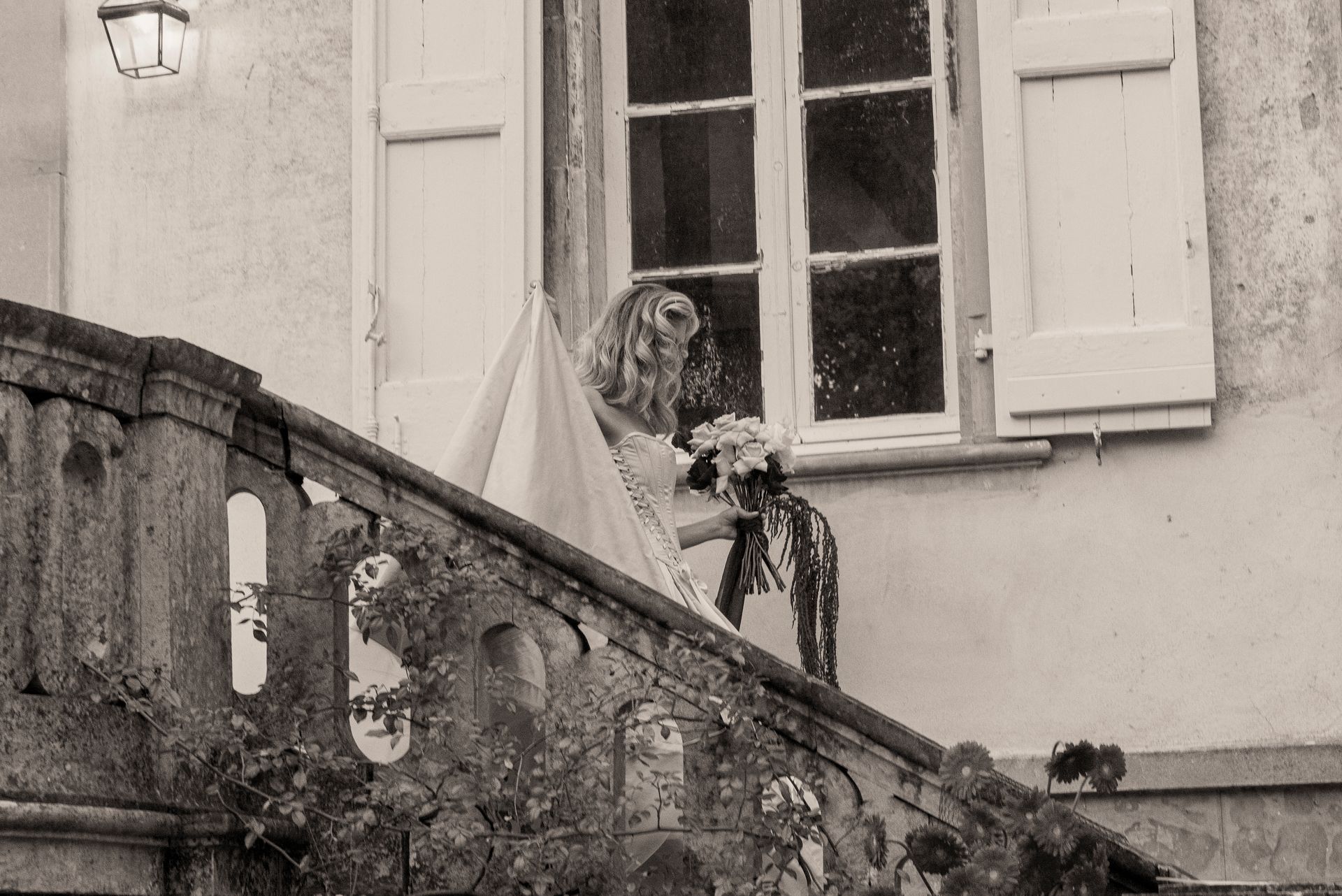 Bride on stone staircase holding bouquet, near window with closed shutters.
