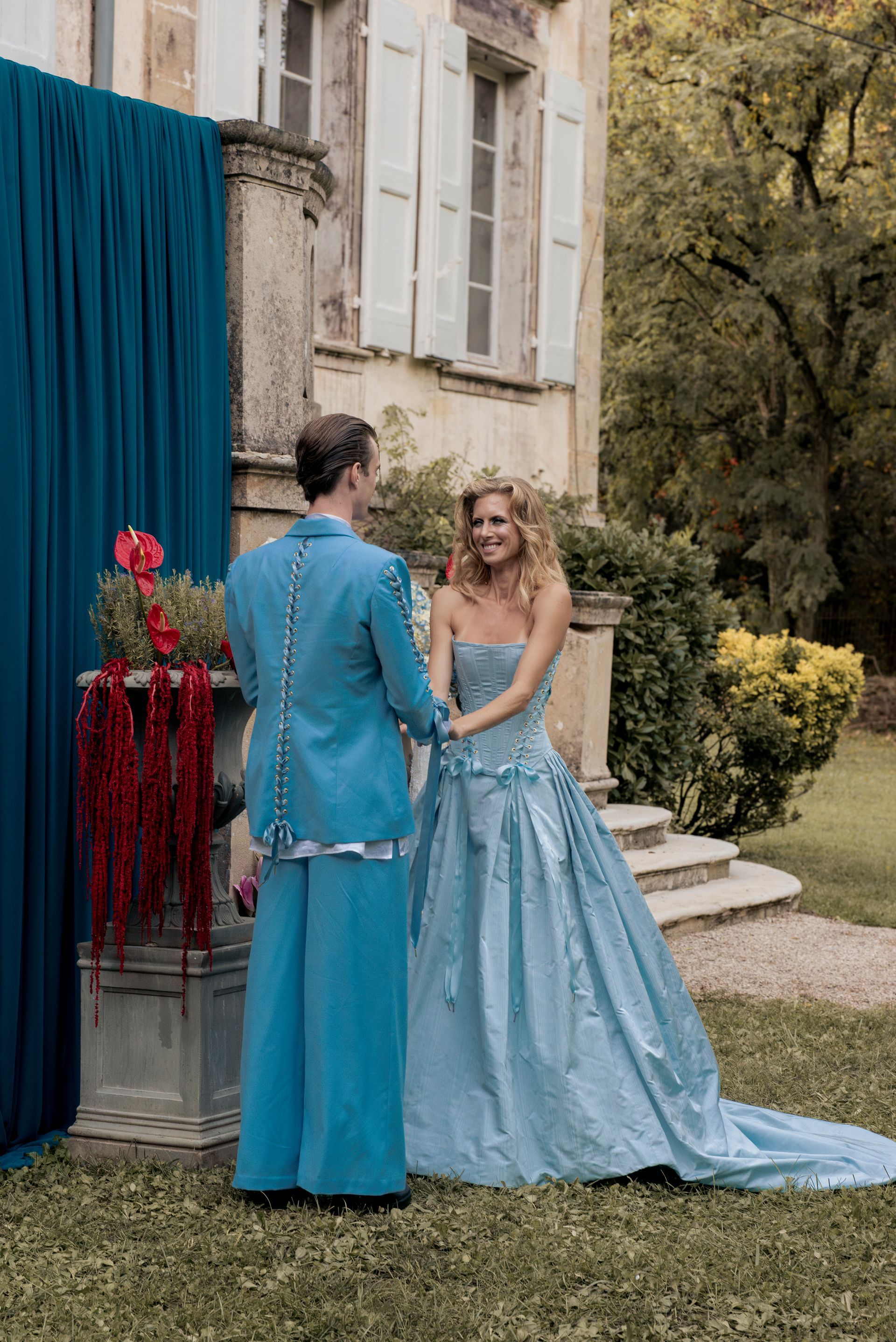 Couple in light blue formal wear, holding hands outside a mansion.
