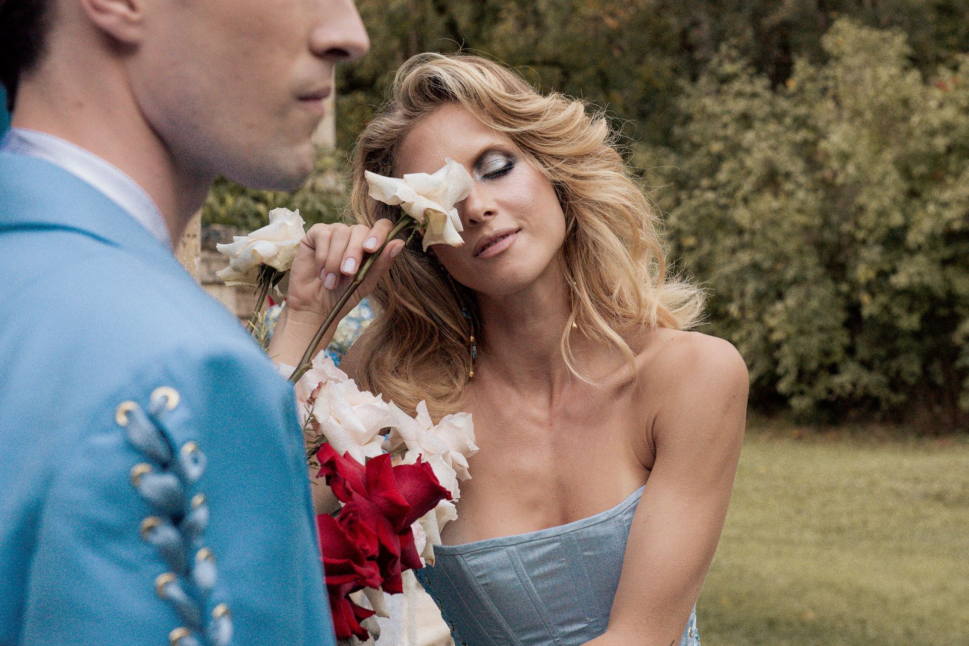 Woman holding roses to her face, eyes closed. Man in blue suit stands beside her outdoors.