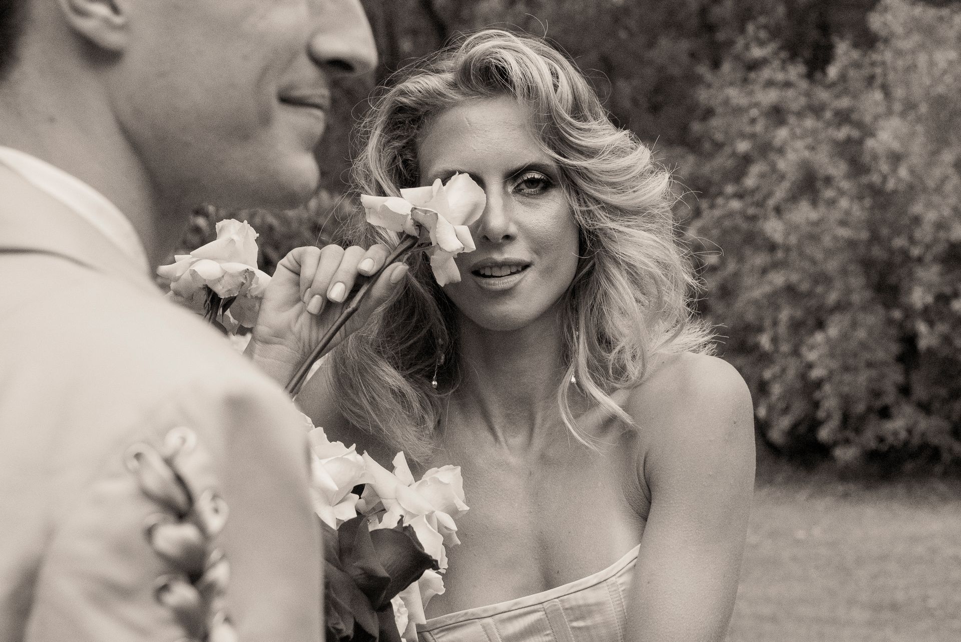 Woman holding flower to eye, smiling. Man in background. Outdoor setting.