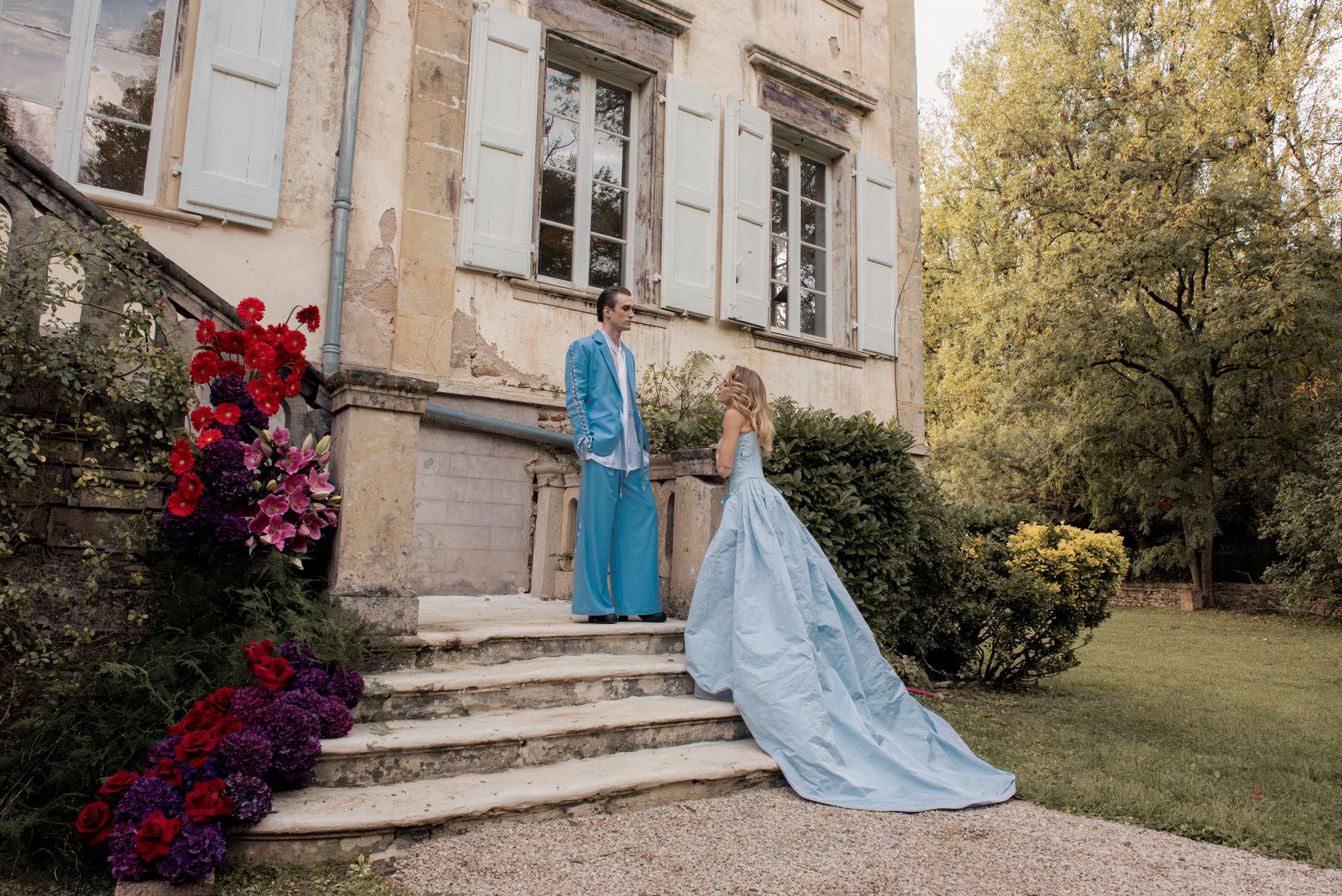 Couple on steps of stone building; woman in blue gown, man in blue suit; flowers.