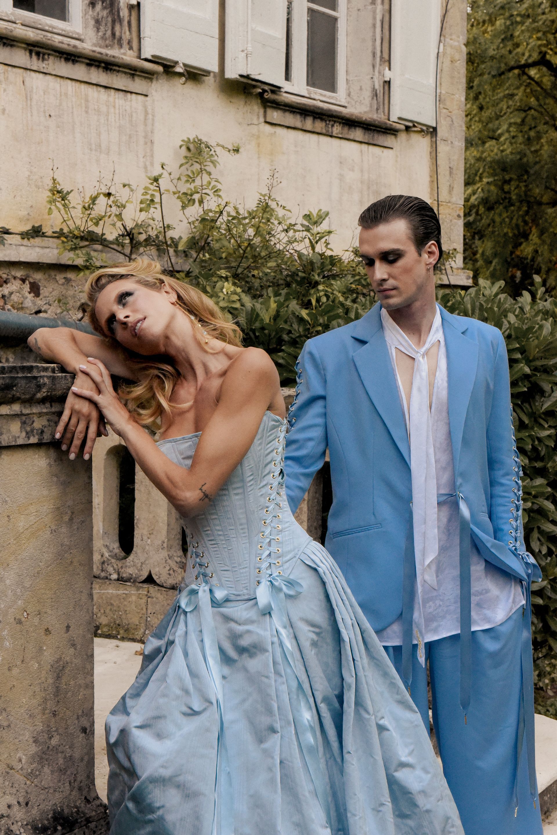 Woman in blue dress leans on stone railing; man in blue suit looks at her. Outdoors.