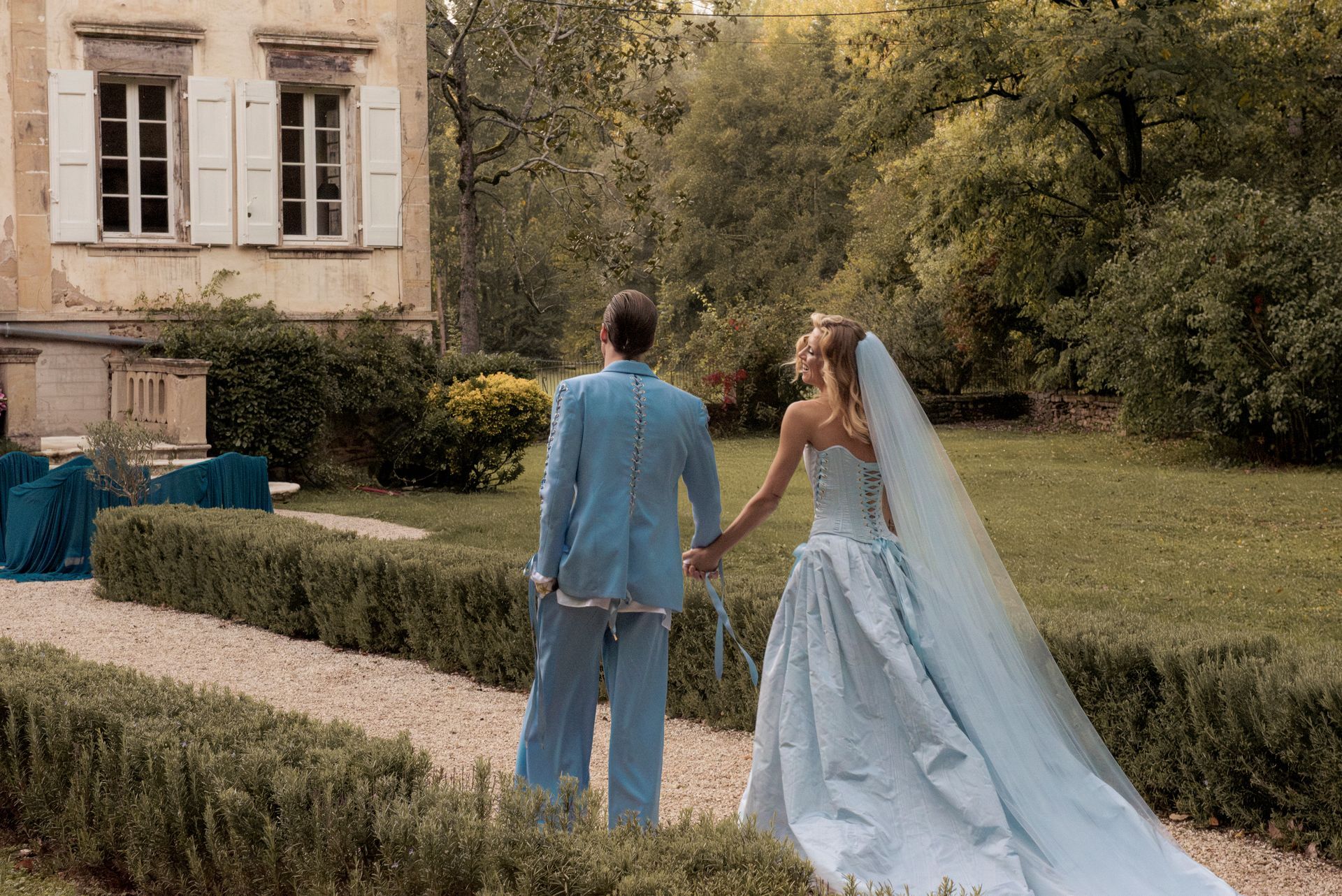Couple in blue wedding attire walk away from camera, holding hands, in a garden.