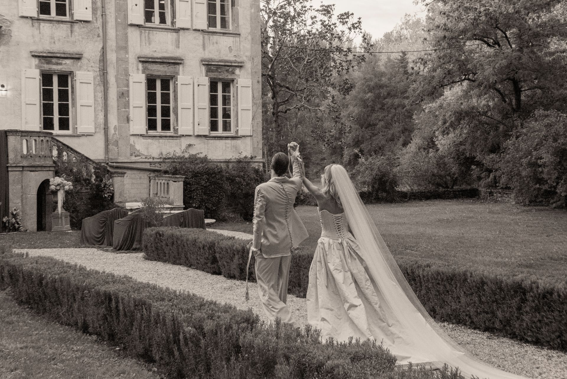 Newlyweds walking away from camera, towards castle. Bride in gown with veil, groom in suit, on path.
