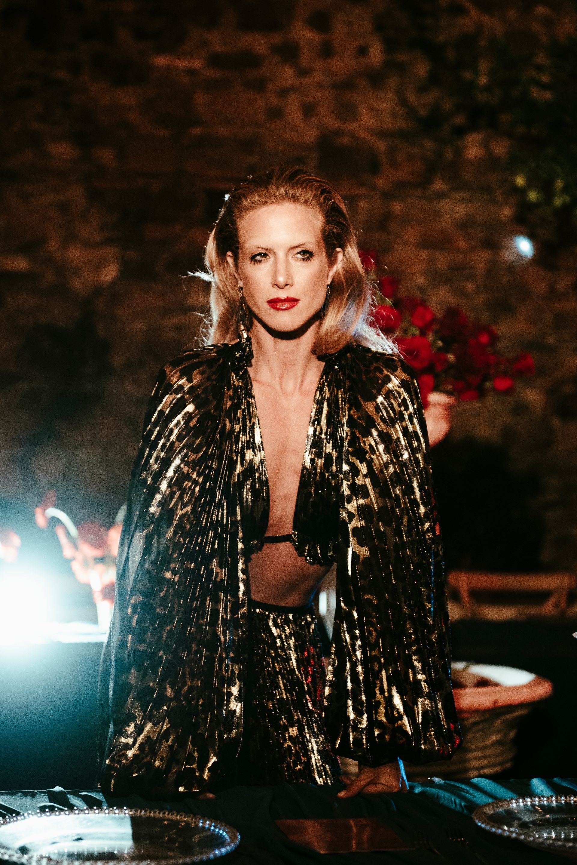 Woman in gold sequined cape, red lipstick, standing by a table, firelight in background.